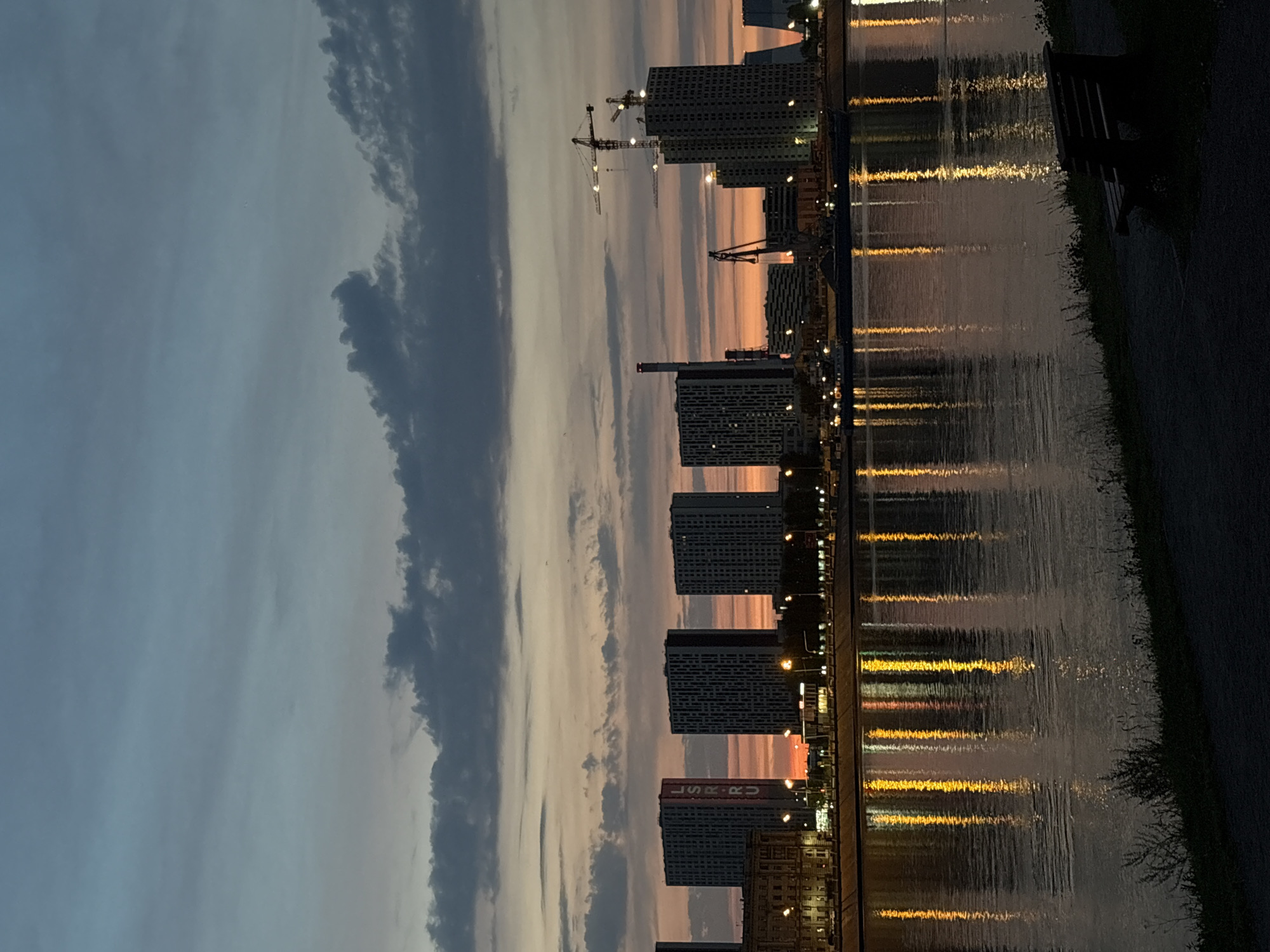 The image depicts a waterfront scene at dusk in Saint Petersburg, Russia, likely along the Neva River. In the foreground, a dark park bench sits on a paved pathway, with sparse vegetation bordering the path. The middle ground features the calm surface of the river, reflecting the lights of the city and the colors of the twilight sky. The city skyline dominates the background, characterized by modern apartment buildings and construction cranes, indicating ongoing development. Notably, one tall building displays the "LSR.RU" logo. The sky is a gradient of deep blues and purples, transitioning to hues of orange and pink near the horizon, with scattered dark clouds. The overall atmosphere is serene and urban.