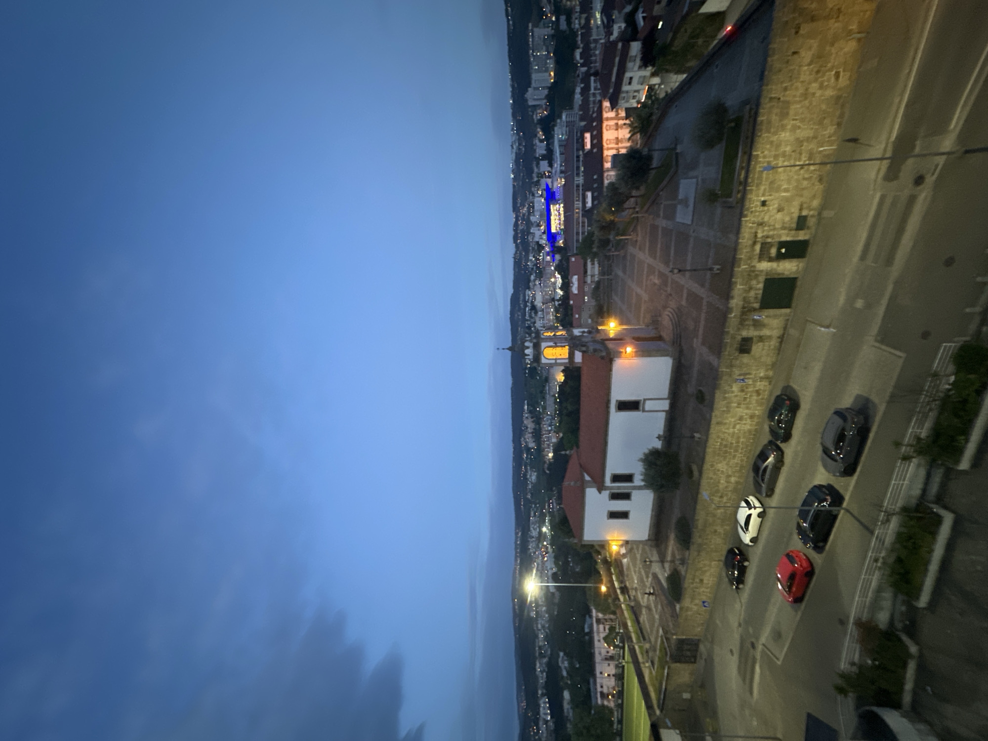 The image depicts an elevated view of Vila Real, Portugal at dusk. The sky is a gradient of blue, transitioning from light to dark. The urban landscape is visible, featuring buildings, streets, and green areas. A prominent white building with a red roof and a lit steeple stands out. A parking area alongside a road contains several parked cars of various colors. Street lights illuminate the scene. The image captures a sense of urban tranquility.