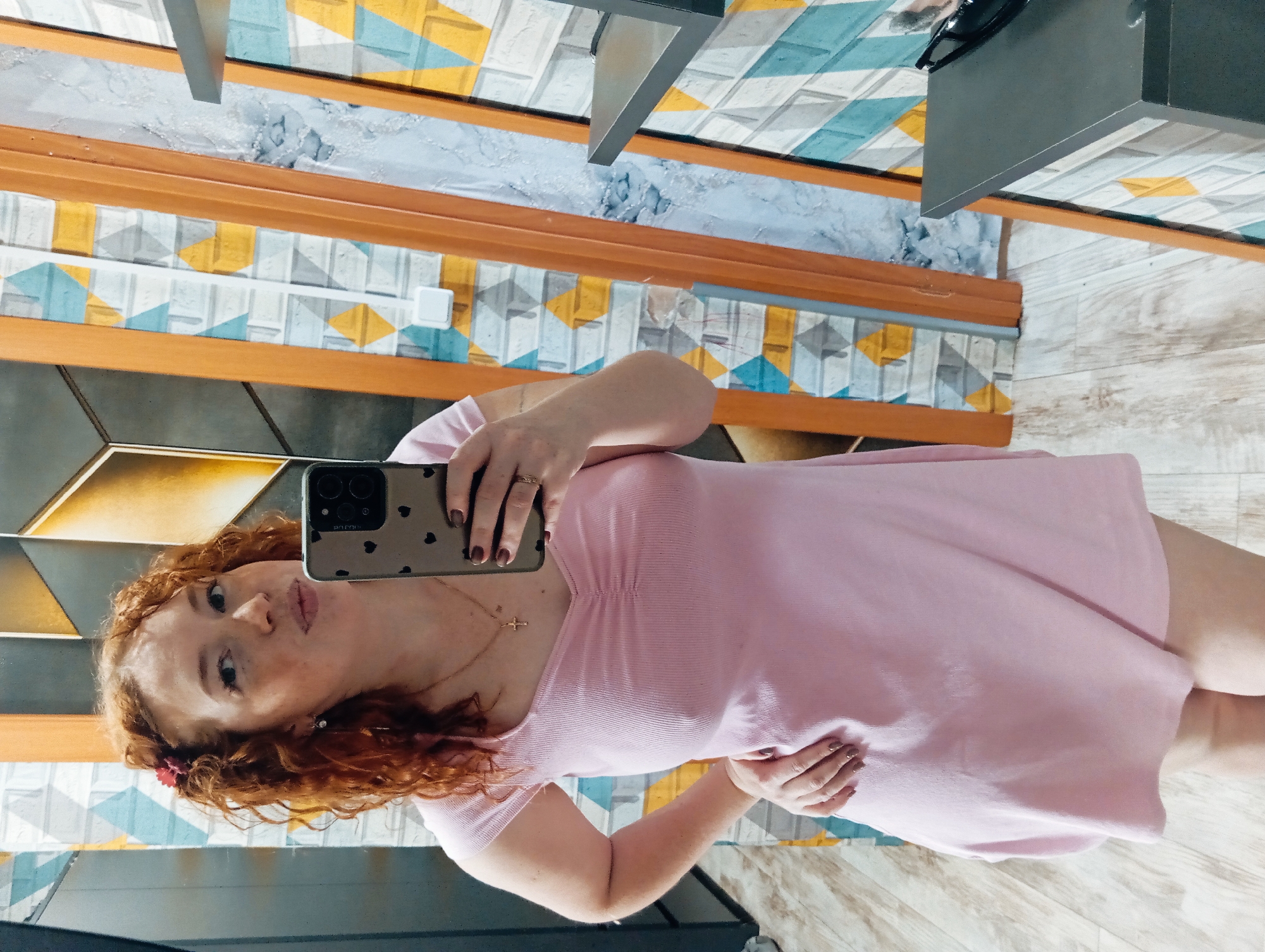 A young woman with wavy auburn hair is captured in a mirror selfie, seen from approximately the waist up. She is wearing a light pink, short-sleeved top or dress. Her head is tilted slightly, and she has a relaxed expression with a subtle pout. A small reddish flower or clip is adorned in her hair on the left side of her head. Around her neck, she wears a thin gold-colored chain with a small cross pendant.
She holds a smartphone in her right hand, which is visible in the reflection. The phone has a light-colored case adorned with small black heart patterns. The text "PRO" is visible on the phone case below the camera module, and some fainter, smaller text like "2mm" is discernible near the camera lenses. Her fingernails are painted a dark plum or brownish-purple color, and she wears a ring on her right ring finger. Her left hand rests casually near her hip or stomach.
The background features a decorative wall with geometric patterns, including squares, diamonds, and triangles, rendered in shades of yellow, light blue, grey, and white. These patterned sections are framed by thick, light brown wooden trim. A white rectangular electrical outlet is visible on one of the patterned wall sections. Above the patterned wall, a dark grey, angular fixture or shelf is partially visible. The lower part of the image shows a light wood-patterned floor. The indoor setting, with its distinct decor and mirror, suggests a personal space or a well-decorated room in a building in Yurgamysh, Russia. The lighting indicates an indoor environment.