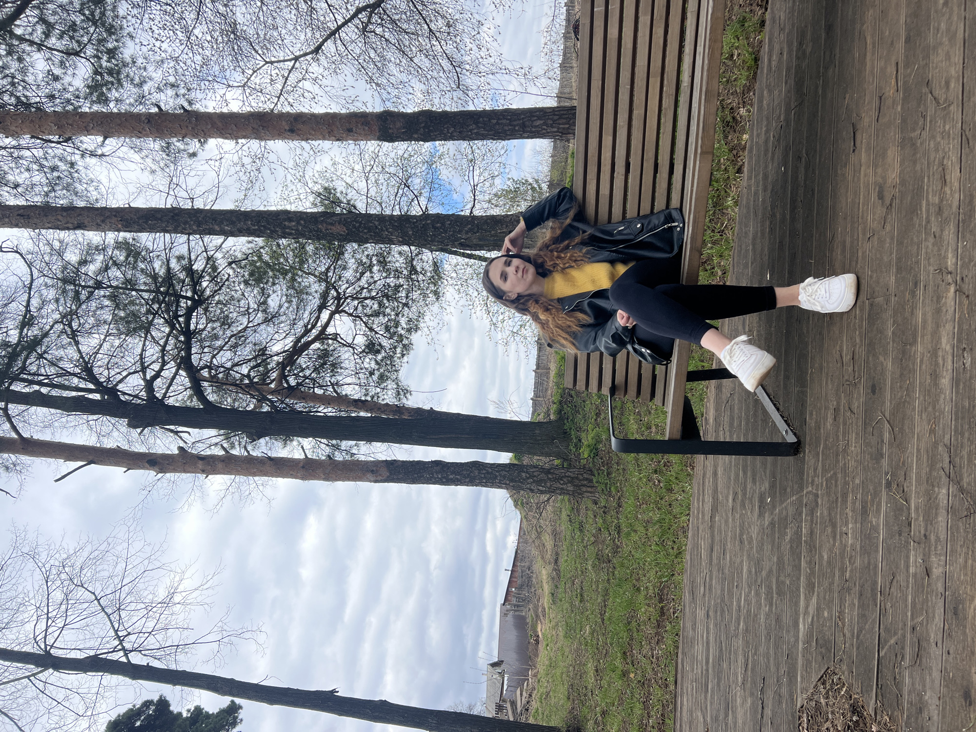 The image captures a young woman lying on her back on a wooden park bench, looking towards the camera. She has long, wavy brown hair and is dressed in a bright yellow top, a black leather jacket, black leggings, and white sneakers. Her left arm is bent, with her hand near her head, and her right arm rests across her body.
The bench is made of wooden slats and has black metal supports. It rests on a dark wooden planked surface, possibly a boardwalk or decking. To the left of the boardwalk, patches of green grass and brown earth are visible.
The setting is outdoors, likely a park or wooded area in Votkinsk, Russia. Several tall tree trunks, appearing to be pines or similar conifers, are prominent in the middle ground, extending vertically from the bottom to the top of the frame. Many of the trees have sparse, bare branches, suggesting either late winter or early spring. The sky is overcast with a mix of light and darker grey clouds. In the far background, behind the trees and a grassy slope, some distant buildings and structures can be faintly seen. The lighting is diffused, consistent with an overcast daytime. No visible text is present in the image.