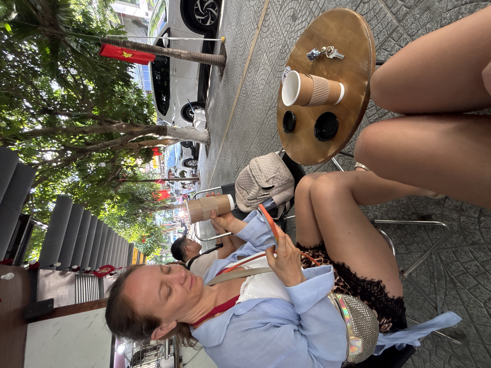 The image depicts an outdoor cafe setting on a patterned gray tiled sidewalk in Đà Nẵng, Vietnam, identified by multiple Vietnamese flags (red with a yellow star) displayed along the street. In the left foreground, a woman with light brown hair, wearing a light blue long-sleeved shirt over a white lace-trimmed top and leopard-print shorts with black lace trim, is seated on a metal chair. She holds an orange-cased smartphone in her left hand and a brown paper coffee cup with a white lid in her right hand, her gaze directed at the phone. A silver iridescent fanny pack is visible across her chest.
Adjacent to her, a small, round, light brown wooden table displays another identical brown paper coffee cup, a separate black lid, and a set of keys with a blue and silver charm. The upper thighs and knees of a second person, presumably the photographer, are visible in the lower right corner, indicating their presence at the table. To the left of the table, a quilted beige backpack rests on a chair.
In the mid-ground, a man with dark hair and glasses, wearing a light-colored t-shirt, is seated further back, looking to his right. The background features a paved sidewalk with a yellow painted line, bordering a street with several white SUVs, including a Toyota Corolla Cross, parked parallel to the curb. Multiple trees with dense green foliage and some with white string lights provide shade. An overhead dark gray retractable awning with horizontal slats and red decorative tassels is visible. The scene is illuminated by bright daylight.