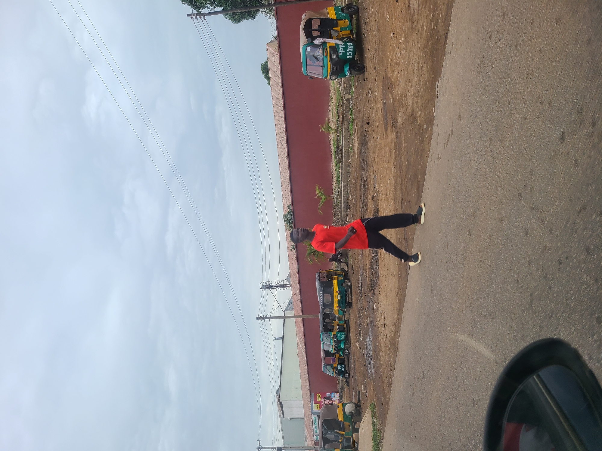 The image shows a street in an urban environment, possibly in the city of Unknown, Unknown. The view is captured from within a vehicle, indicated by the partial view of a car's interior on the bottom right. The road stretches out into the distance, with an individual wearing a bright orange shirt and dark pants seemingly walking or running along the side of the road. Several green and yellow auto-rickshaws (tuk-tuks) are parked alongside a building with a red exterior. Electrical wires are strung overhead, and the sky is overcast with grey clouds. The ground surface near the auto-rickshaws appears to be a mix of dirt and vegetation.