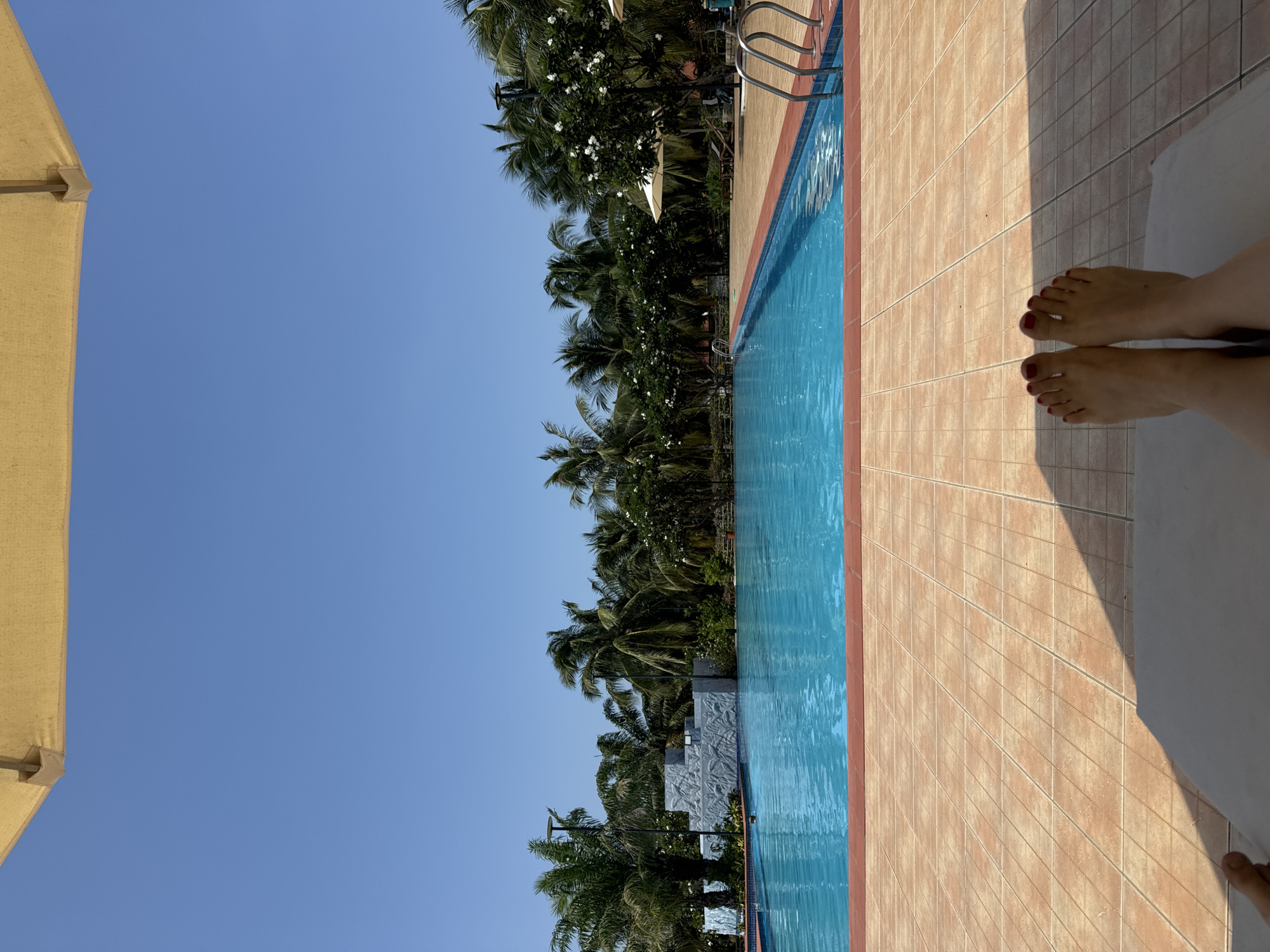 The image shows a resort swimming pool in Phú Thủy, Vietnam, on a sunny day. The pool is large and filled with bright blue water, with a red tile border. Palm trees and other lush green vegetation surround the pool, with a clear blue sky overhead. In the foreground, a person's bare feet are visible, resting on a lounge chair. The feet are adorned with red nail polish. A large yellow umbrella is partially visible at the top of the frame, providing shade. The scene suggests a relaxed vacation atmosphere.