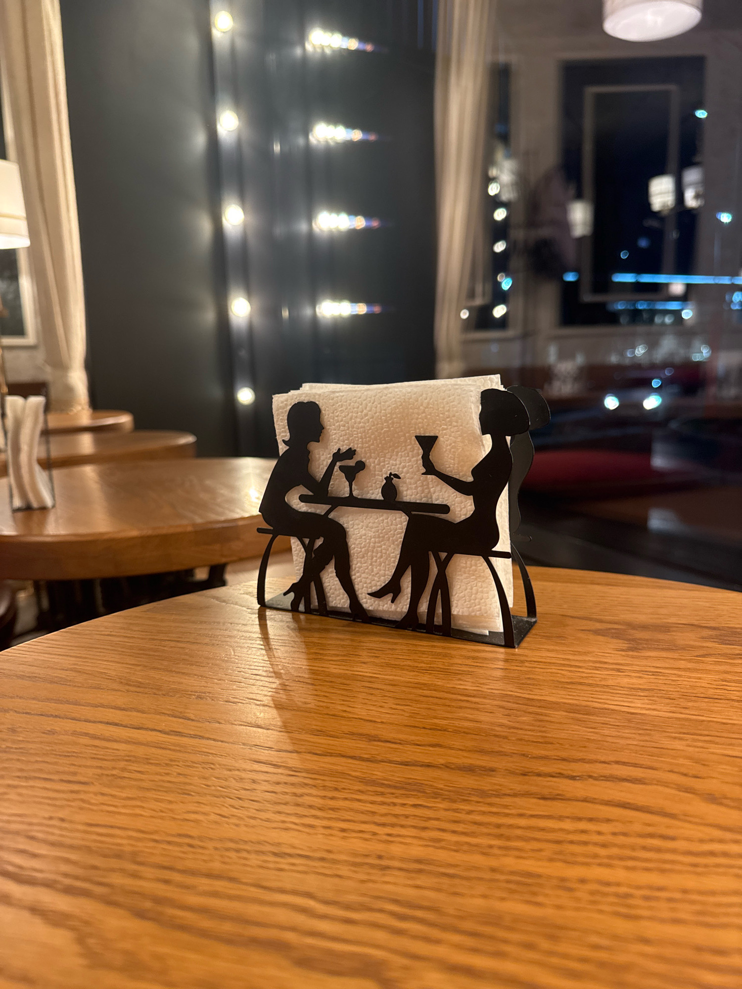 The image depicts a close-up view of a wooden table in what appears to be a cafe or restaurant. In the foreground, a napkin holder is visible. It is designed as a black silhouette of two women sitting at a table, each holding a drink. A stack of white napkins is held within the holder. The background is blurred, showing other wooden tables, soft lighting from what seems to be string lights, and a window with reflections of city lights, suggesting an urban setting. The overall ambiance is dim and cozy. Given the context, this scene could plausibly be located in a cafe in Ufa, Russia, as cafes with such decor are common in urban environments worldwide.
