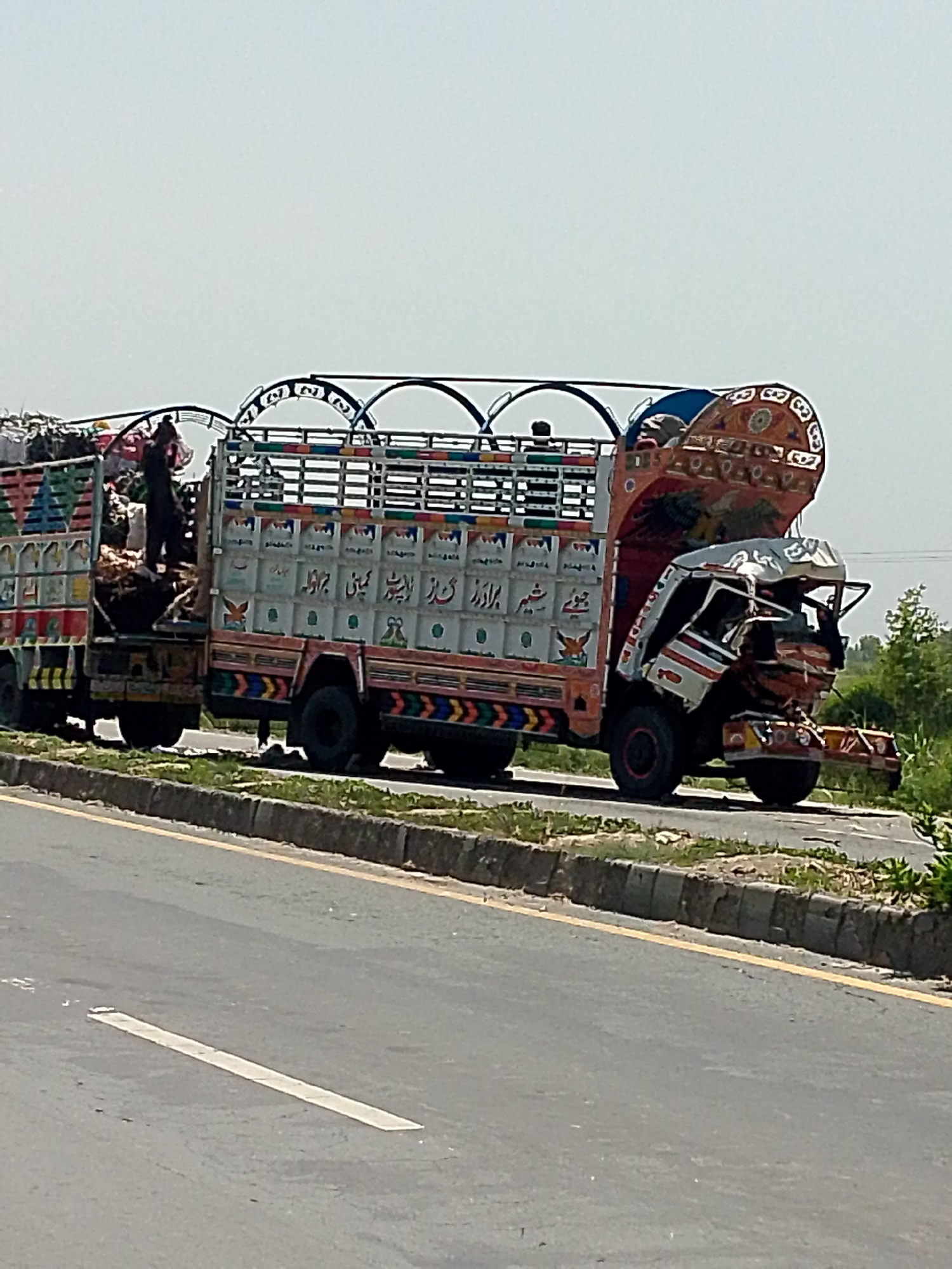The image shows a brightly decorated truck with a damaged front end on the side of a road. A man is standing on top of the cargo in the back of the truck. The road is asphalt with a curb and vegetation on the side. The location is in the city of Unknown, Unknown.