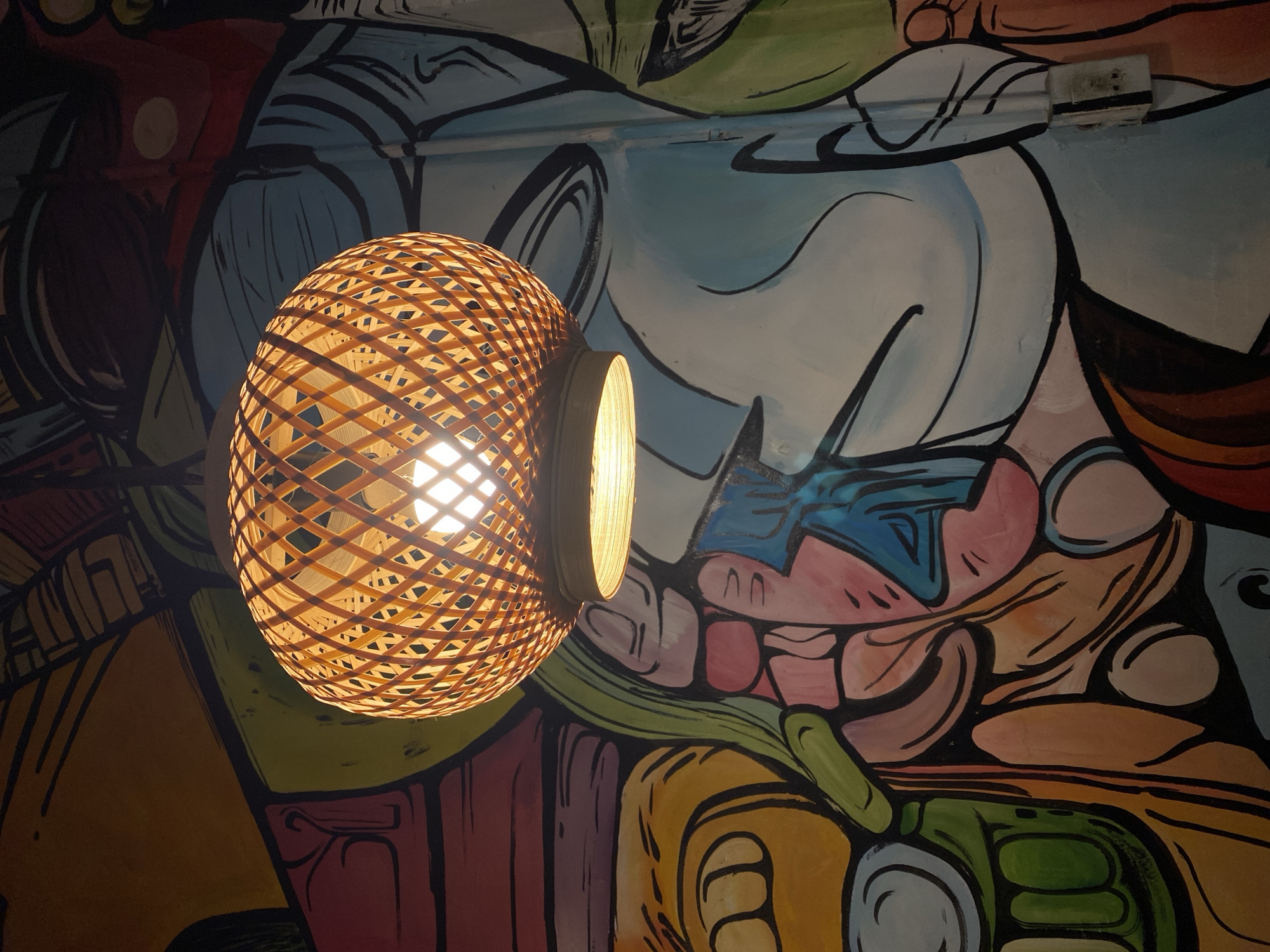 An overhead, eye-level, medium shot captures a woven, circular bamboo pendant light fixture hanging in front of a colorful mural. The light fixture, made of light brown bamboo strips woven in a diamond pattern, emits a warm, yellow glow from an exposed lightbulb within. The mural behind it features bold, abstract shapes and lines in a variety of vibrant colors, including yellow, orange, red, pink, green, blue, and black. The style of the mural suggests a modern, artistic aesthetic. The image is taken from a low angle, looking up at the light fixture and mural. The scene is likely an interior space, possibly a restaurant, cafe, or shop, given the decorative lighting and mural. The location is identified as Đà Nẵng, Vietnam.
