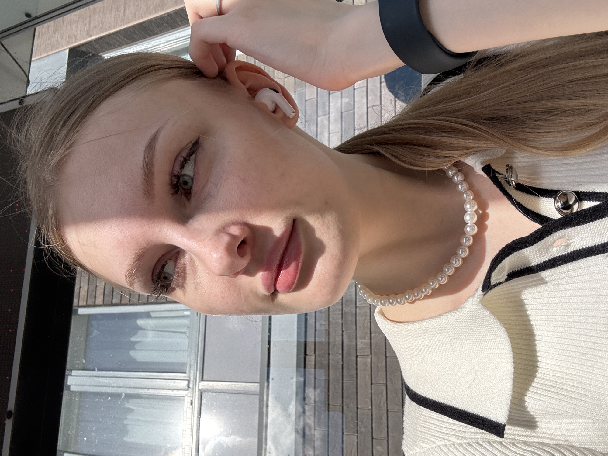 A young woman with light brown hair is captured in a close-up, outdoor, profile shot. She is wearing a white ribbed sweater with black trim and a pearl necklace. A single white wireless earbud is in her right ear, and her right hand is near her ear, with a ring on her index finger. Her left hand is not visible. The background consists of a brick wall and a window with white curtains. The lighting suggests it is daytime and sunny, with strong shadows casting across her face and the environment. The image does not contain any elements that specifically indicate the location is the city of Perm, Russia.