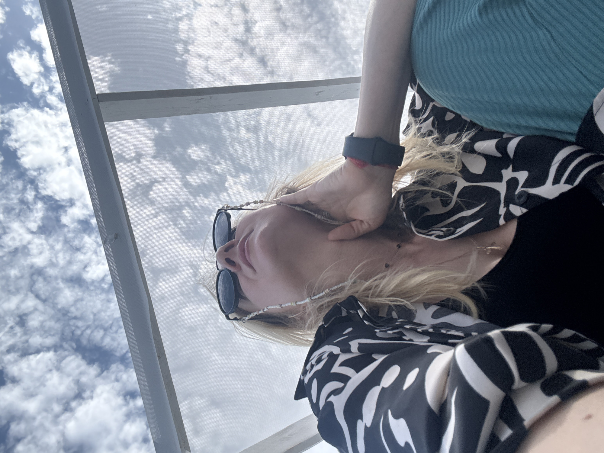 A low-angle shot depicts a person wearing sunglasses and a patterned shirt, possibly standing under a structure with a screen roof, as suggested by the view of the sky and clouds filtered through the screen. The person is wearing a black fitness tracker on their wrist and is positioned in a way that the camera is looking up at them. The location appears to be outdoors due to the presence of the sky. The city is not discernible from the image.