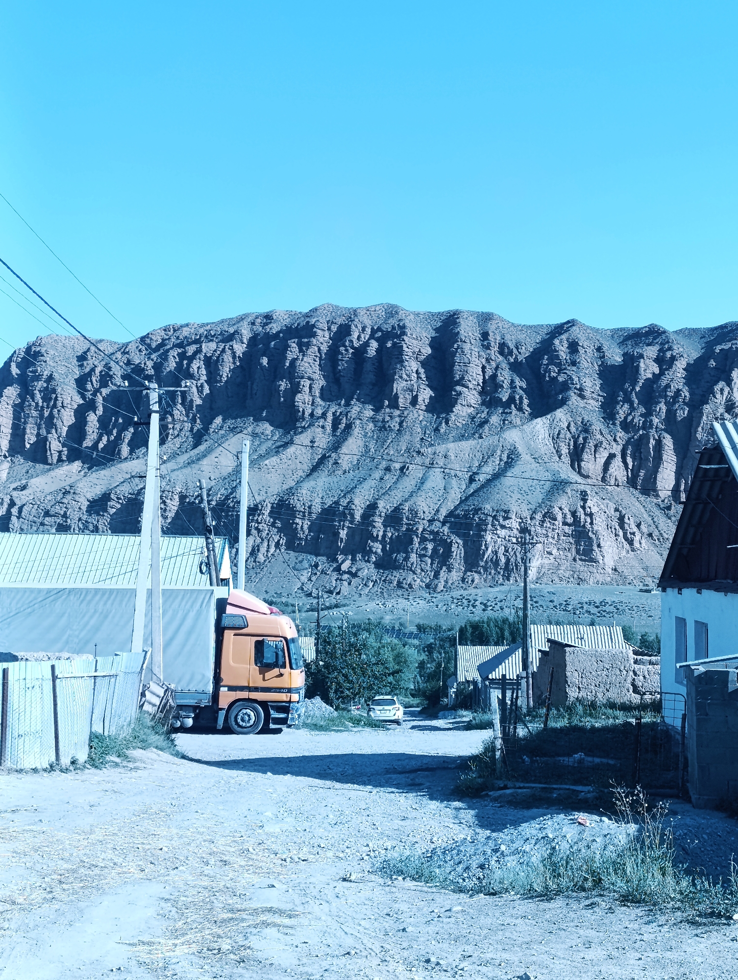 A bright, clear sky dominates the upper portion of the image, with a range of rugged, layered mountains stretching across the horizon. In the foreground, a dirt road curves through a rural village. To the left, a light-colored truck with an orange cab is parked on the side of the road, next to a fence and a building with a light-colored tarp. Several poles with wires run across the middle ground. Further back, a few small, modest houses with corrugated metal roofs are visible, and a white car is parked on the road. The overall scene depicts a quiet, sun-drenched day in a mountainous, semi-arid region, likely a village on the outskirts of Naryn City, Kyrgyzstan.