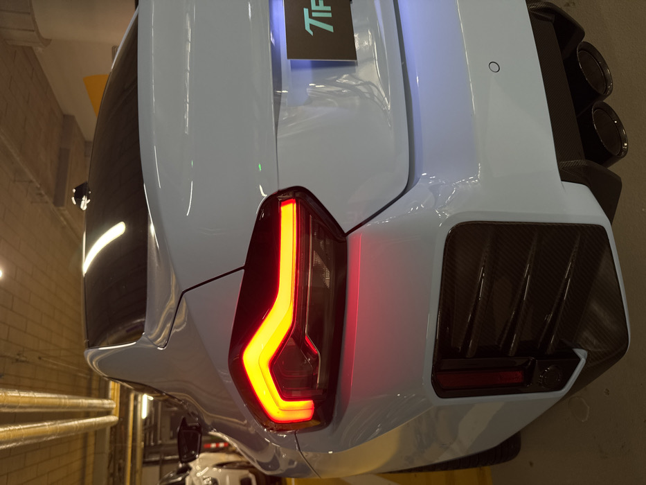 The image depicts the rear three-quarter view of a light blue high-performance sports coupe, likely a modern BMW G87 M2, situated in an indoor parking garage or detailing facility at Tiff Detailing, plac Konstytucji 5, Warsaw, Poland. The vehicle's left taillight is illuminated with distinct red and amber LED elements. The rear bumper features a prominent black carbon-fiber patterned diffuser assembly, including vertical vents on the left side and four black exhaust tips on the right. A single black circular parking sensor is visible on the light blue bumper. Below the trunk lid, a black rectangular display panel with a partially visible, illuminated light blue "Tiff" logo is present. The car has a tinted rear window and a black shark fin antenna on its roof. The environment consists of a concrete ceiling with exposed silver ventilation ducts and warm yellow recessed lighting. The floor is dark. A partial view of a white vehicle is visible in the background on the far left. No individuals are present in the scene, and no specific actions or interactions are depicted.