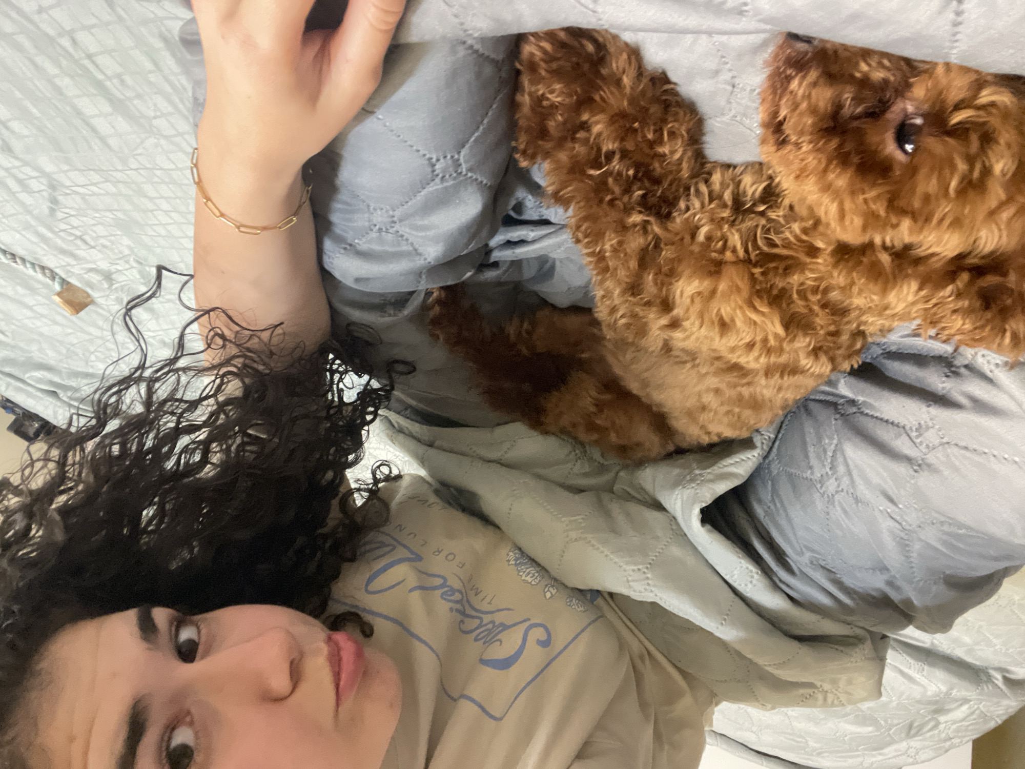 A young woman with dark curly hair is lying in bed next to a brown poodle. The woman is wearing a beige t-shirt with blue lettering. She has a gold bracelet on her left wrist. The bed has light blue and gray quilted covers. The location is unknown and there is no information to indicate the image was taken in Sofia, Bulgaria.