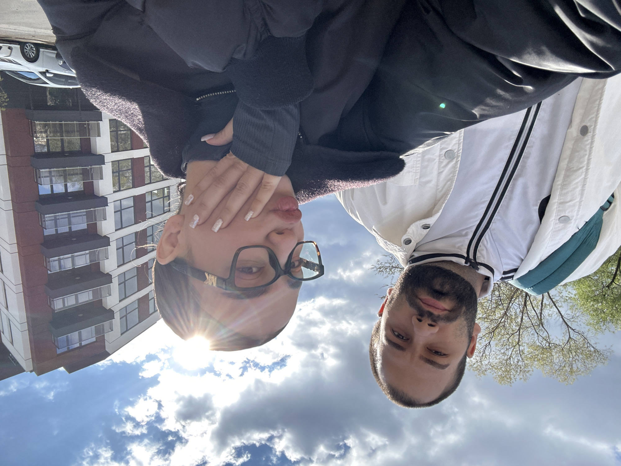 Two individuals are positioned closely together, with the woman on the left and the man on the right, looking down towards the camera. The woman is wearing glasses and a dark jacket, partially covering her face with her hand, and is positioned against a backdrop of a modern building with repetitive window patterns. The man is wearing a white jacket with black trim and a green detail, with a backdrop of trees and a cloudy sky. The image appears to be taken from below, tilting the subjects and the background. Based on the building's architectural style, it is plausible that this picture was taken in Sofia, Bulgaria, but there is no explicit proof of the location.