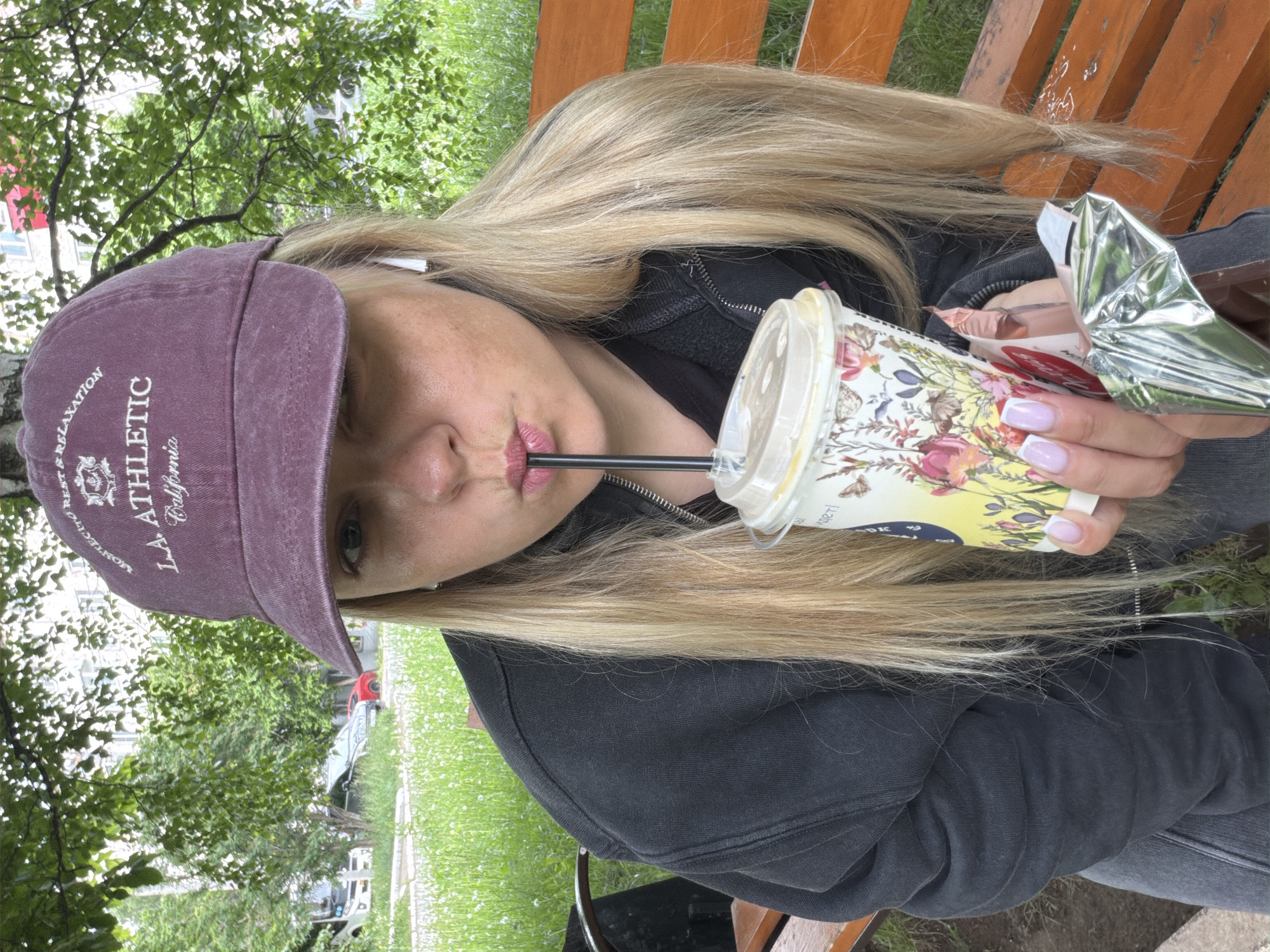 A woman with long, blonde hair is captured in a close-up, outdoors shot, seated on a wooden slatted bench. The image is rotated, showing her looking up towards the camera. She wears a dark purple baseball cap embroidered with "REST & RELAXATION," "LA ATHLETIC," and partially "California," along with an emblem. She is dressed in a black zip-up hoodie over a dark top and has earbuds in. Her lips are painted with light pink lipstick, and her fingernails feature a light pink or purple French manicure.
The woman is engaged in a casual activity, sipping from a clear plastic cup with a black straw. The cup is adorned with a colorful floral and botanical pattern, including butterflies and bees. Visible Cyrillic text on the cup reads "РК" and "1932-Т." She is also holding a partially opened snack or candy bar wrapped in shiny silver foil, revealing a pink inner wrapper.
The scene is set in an outdoor green space, likely a park or recreational area in Votkinsk, Russia. The background features lush green trees and grass, with dappled natural light suggesting it is daytime. Some faint structures or elements of a distant background are visible through the foliage. The overall atmosphere is relaxed and informal.