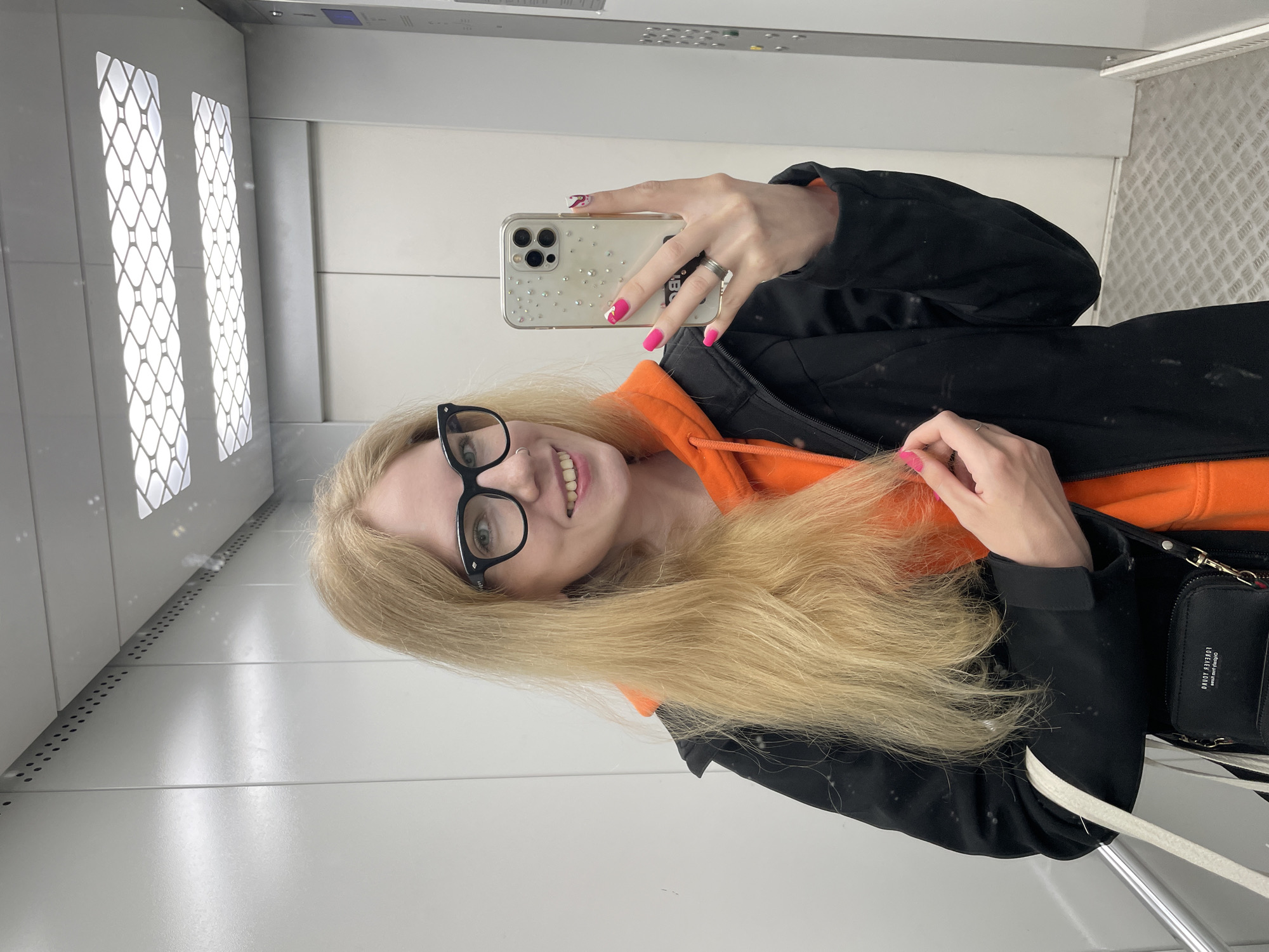 A bright, vertically oriented selfie captures a young woman inside an elevator. She has long, straight blonde hair, black-framed glasses, and a cheerful smile. She is wearing a vibrant orange hooded sweatshirt beneath a black zip-up jacket. Her hands are visible, with fingers adorned with bright pink nail polish and a silver ring on her left ring finger. She holds an iPhone, identifiable by its triple camera array, housed in a clear case decorated with small, sparkling rhinestones. A small black purse with metallic accents and the text "LOVELY KRONI" is visible on her right side.
The setting is an elevator interior, characterized by light-colored, reflective walls. On the left wall, two tall, illuminated panels feature a distinctive white, lattice-like pattern. The floor has a textured, grey metallic surface. The bright, artificial lighting suggests an indoor public space. The woman's smiling expression indicates a positive mood. The image captures a casual moment of a selfie being taken. The external location context provided is Izhevsk, Russia.