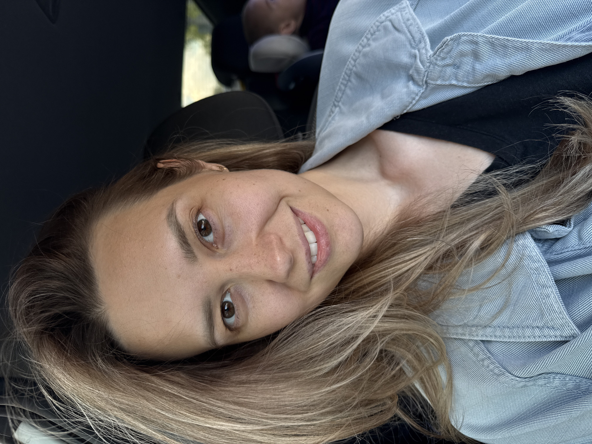 The image displays a close-up portrait of a woman, captured sideways, as if the camera was rotated or she is leaning her head. She has light skin with subtle freckles across her nose and cheeks, and warm brown eyes. Her long, light brown hair with blonde highlights cascades downwards towards the right side of the frame, partially covering a light blue denim jacket worn over a black top. A pleasant and gentle smile is visible on her face, revealing her upper teeth.
The background is dark and blurred, indicating the interior of a car. Towards the upper right corner of the frame (considering the sideways orientation), a partial view of a baby or small child in a car seat can be seen. The natural and even lighting suggests the photo was taken during the daytime. The woman's expression conveys a sense of happiness and relaxation. No specific landmarks or identifying features are visible within the car or through the windows that would indicate the location as Chaikovsky, Russia. There is no visible text in the image.