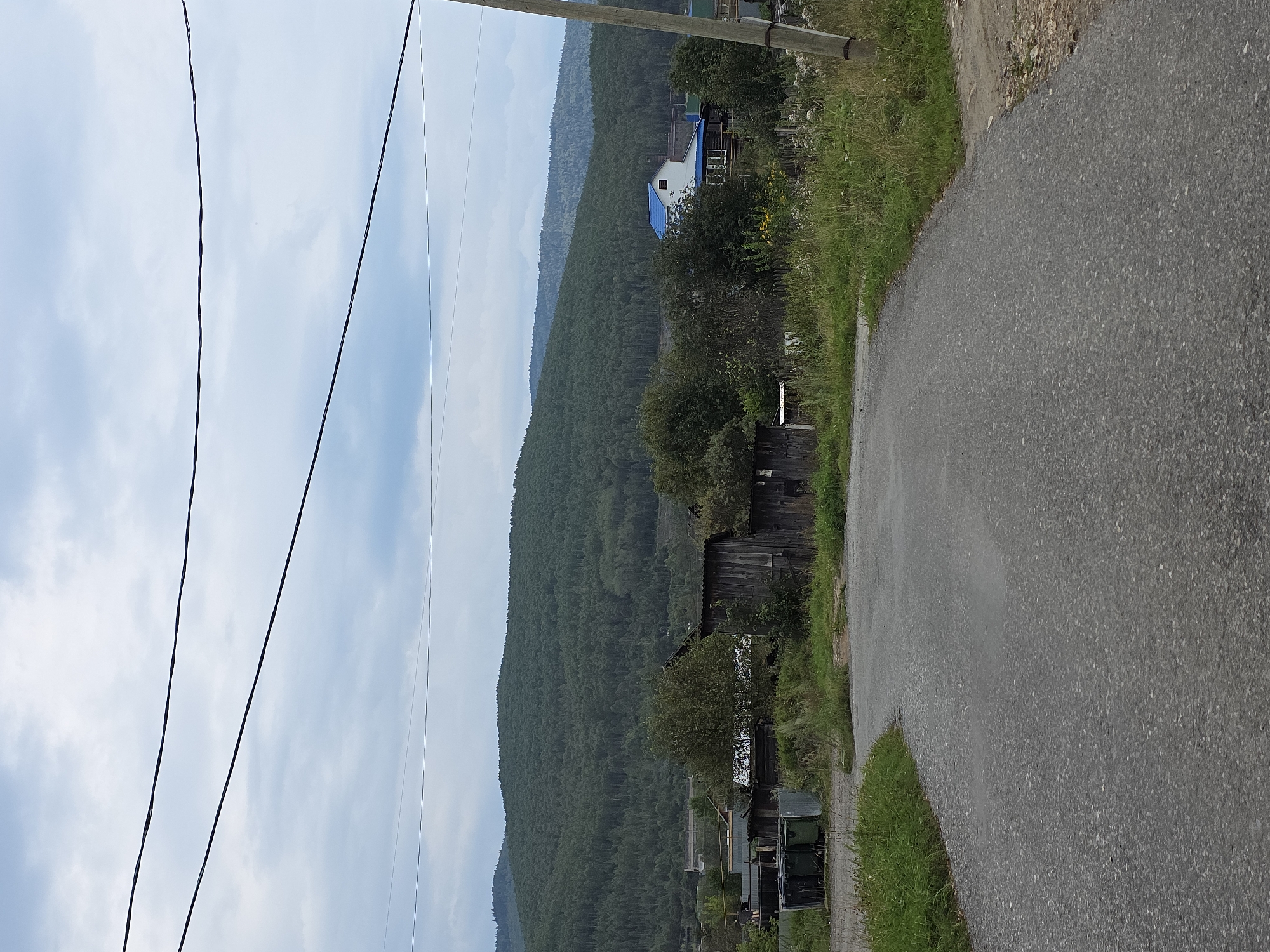 This image depicts a rural scene in the city of Magnitka, Russia. A paved road curves through the foreground, leading towards a cluster of wooden buildings and a modern house with a blue roof nestled among trees. In the background, a densely forested hill dominates the landscape under a cloudy sky. Power lines crisscross the upper portion of the image. The scene appears to be an overview of a small settlement within a natural environment.