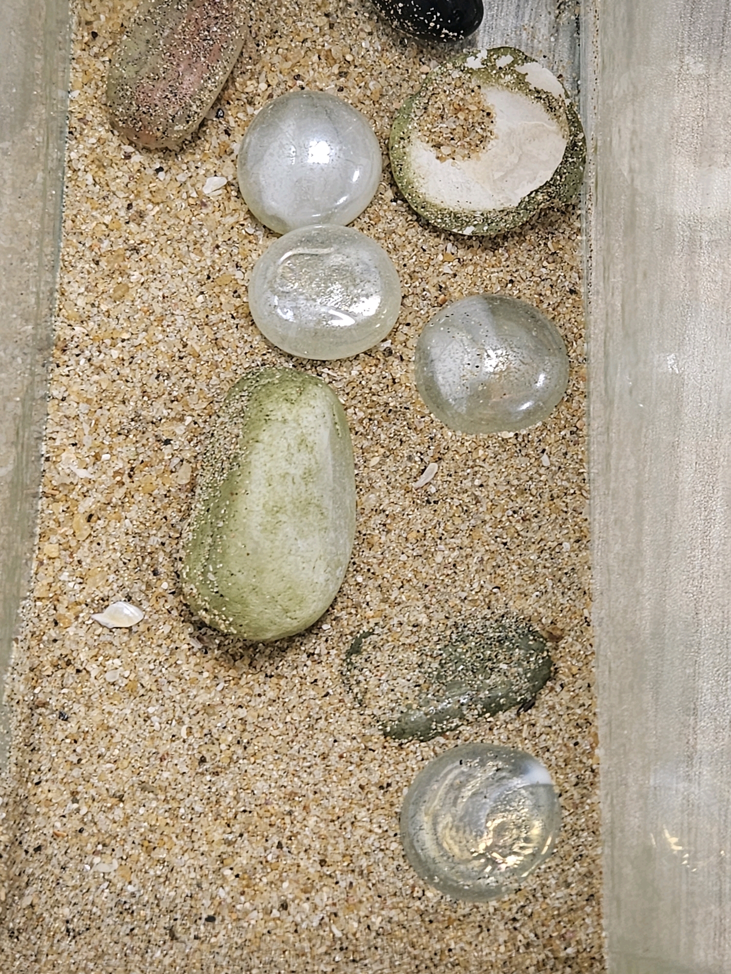 The image shows a close-up, top-down view of a shallow container filled with sand and various decorative objects, including smooth, rounded stones and glass marbles. The sand appears to be composed of fine grains of varying colors, predominantly beige and light brown, with some darker flecks. Several objects are partially or fully embedded in the sand.
On the left side of the frame, there is an elongated, oval-shaped stone with a rough, speckled surface, appearing pinkish-brown. Above and to the right of this stone are three clear glass marbles, two of which are spherical and one slightly flattened. These marbles have a pearlescent sheen and appear to contain small air bubbles or internal imperfections.
Below the marbles, towards the center of the image, is a single, elongated, smooth stone with a pale green hue, mottled with lighter shades. To the right of this green stone, a small, irregularly shaped, dark green object is visible, also partially covered in sand.
In the upper right quadrant, there is a broken shell fragment or a similar calcified object, with a rough, sandy exterior and a white, crystalline interior. A small, dark, spherical object, possibly another stone or marble, is partially visible in the very top edge of the frame, to the left of the broken shell.
A small, white, shell-like object is situated in the sand below the green stone, near the bottom left of the frame. At the bottom center of the image, another clear glass marble with swirling patterns is visible.
The container itself appears to have clear, transparent walls on the left and right edges of the frame, suggesting it might be a display case or part of an aquarium. The lighting is even, illuminating the scene without harsh shadows. The overall impression is of a curated collection of natural and decorative elements within a sandy substrate.
The provided image and context do not contain any information that would allow for the identification of a specific location within the city of Colombo, Sri Lanka.