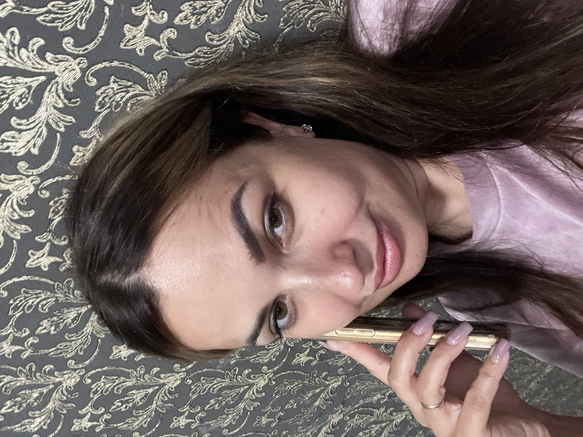 In a close-up shot, a woman is holding a golden smartphone to her right ear, looking towards the camera. She has brown hair and brown eyes, and is wearing a light pink t-shirt and a ring on her left ring finger. Her fingernails are painted a light purple color. Behind her, a dark grey wallpaper with a golden floral pattern is visible. The image appears to be taken indoors.