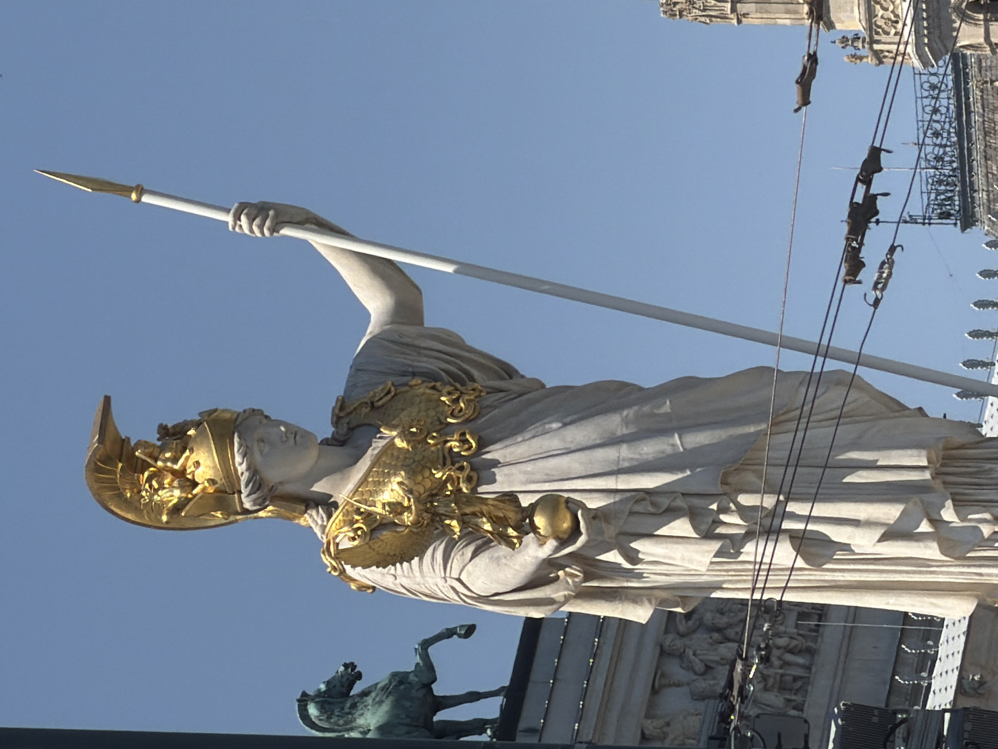 This is a medium shot, eye-level, outdoor, daytime, vertical photograph of the Pallas Athene monument in front of the Austrian Parliament Building in Vienna, Austria. The sky is clear and blue. The Pallas Athene statue, made of marble and gold, stands tall, holding a spear in her right hand and a sphere in her left. She is adorned in a golden helmet and breastplate, with intricate details. To the left of the main statue, a bronze horse statue is visible, partially obscured by the Pallas Athene statue. In the background, the intricate architecture of the Austrian Parliament Building can be seen, along with overhead wires and a section of a building with a spire. The sunlight casts shadows on the statues and buildings, indicating it is a bright, sunny day.