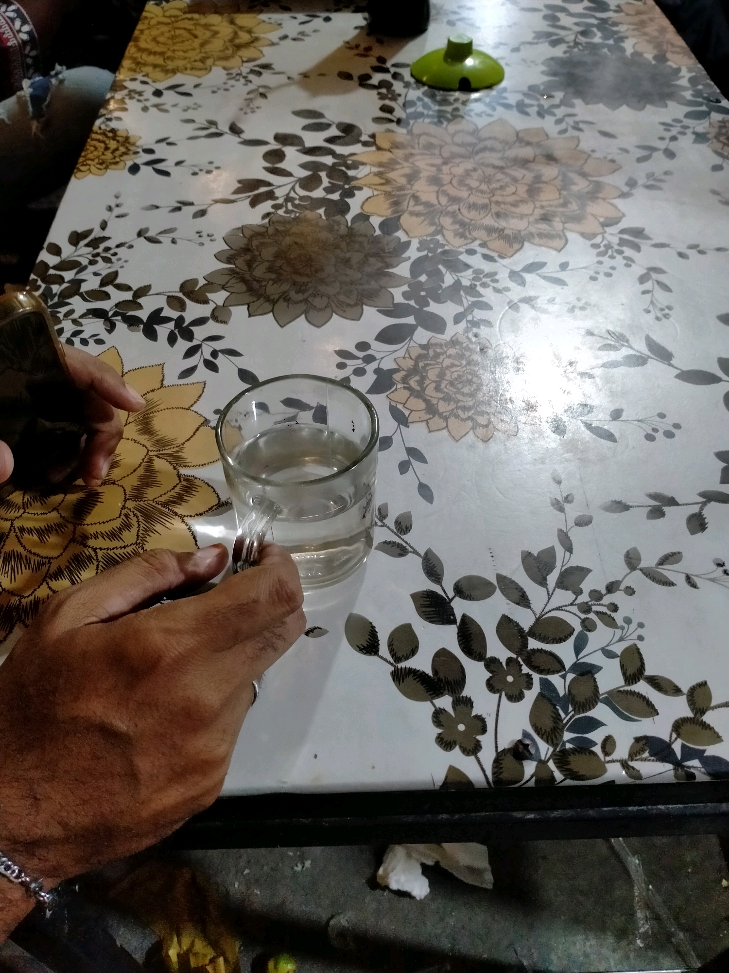 In Ambon, Indonesia, a close-up shot captures a person's hands interacting with a glass of water on a patterned table. The table has a white surface adorned with floral designs in shades of yellow, brown, and gray, featuring large, stylized flowers and smaller leafy branches. A clear glass filled with a clear liquid is positioned on the table, and one hand is seen holding its handle. Another hand is visible to the left of the glass, appearing to be in motion. In the upper right quadrant of the image, a small, green object, possibly a lamp base or a decorative item, rests on the table. The background shows a dark surface below the table, hinting at a floor or lower surface. The lighting appears to be overhead, creating some glare on the table's surface.