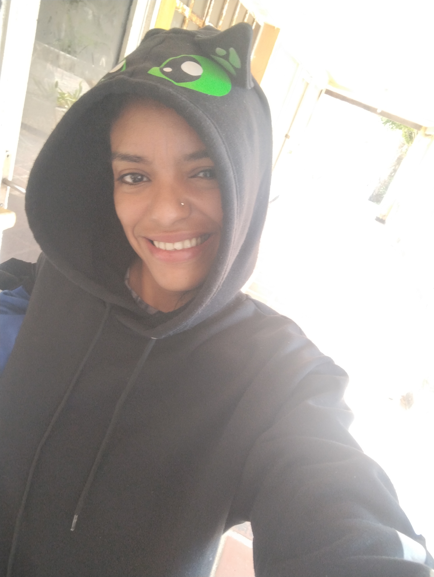 A woman with dark skin and a nose piercing smiles at the camera while wearing a dark hooded sweatshirt with a green and white alien design on the hood. She appears to be indoors, possibly near a window or doorway, as there is bright, diffused light in the background. The environment suggests a well-lit interior space, but specific details about the location in Porlamar, Venezuela are not discernible from the image.