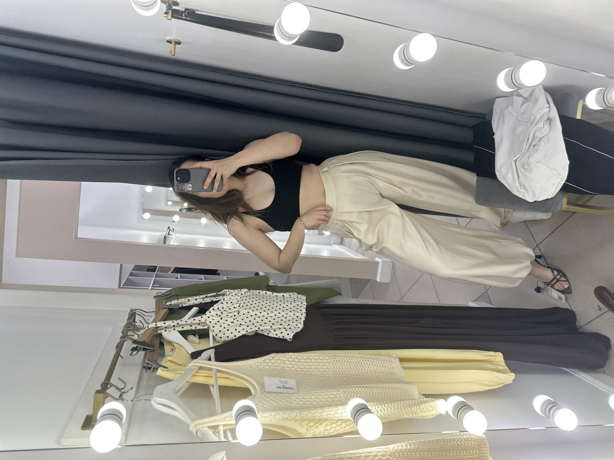 The image captures a woman taking a selfie in a fitting room. She is wearing a black top and light-colored trousers. In the mirror's reflection, clothes hanging on racks are visible, including a white top with black polka dots, a brown dress, and a beige knit top. There is also a pile of white clothes on a seat. The fitting room features a mirror surrounded by light bulbs and a dark gray curtain. Based on the context, this photo may have been taken in a clothing store in Perm, Russia, given the earlier instruction.