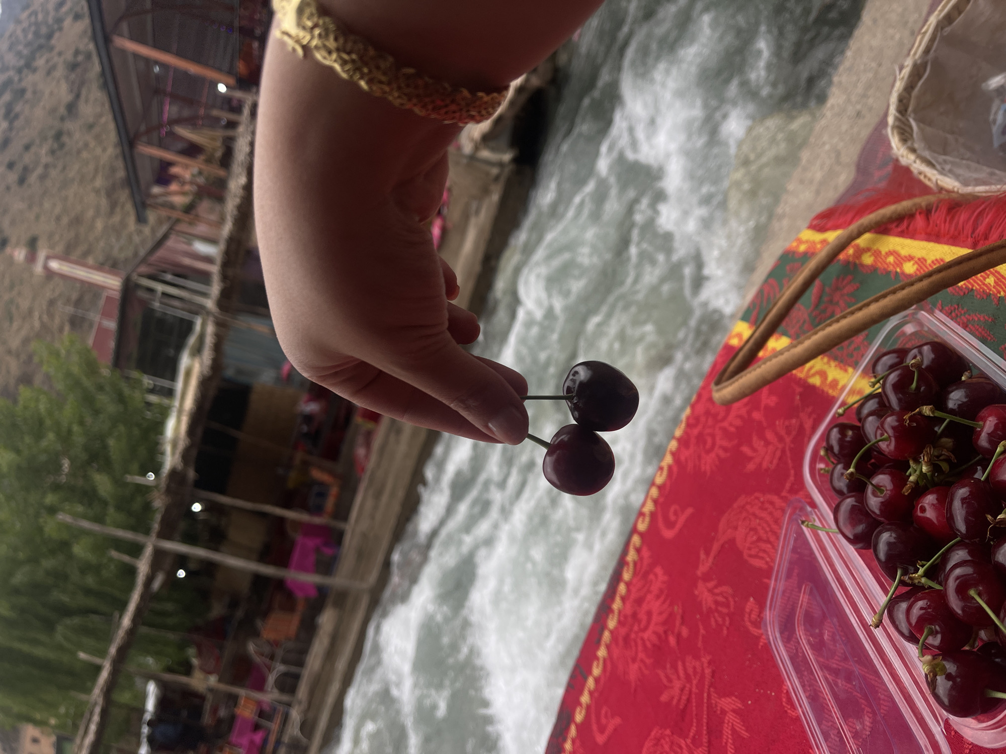 In Asgaour, Morocco, a hand with a gold bracelet holds two cherries over a flowing river. A container full of cherries rests on a red and yellow patterned cloth next to the river. A building with a tower can be seen in the background amongst trees.