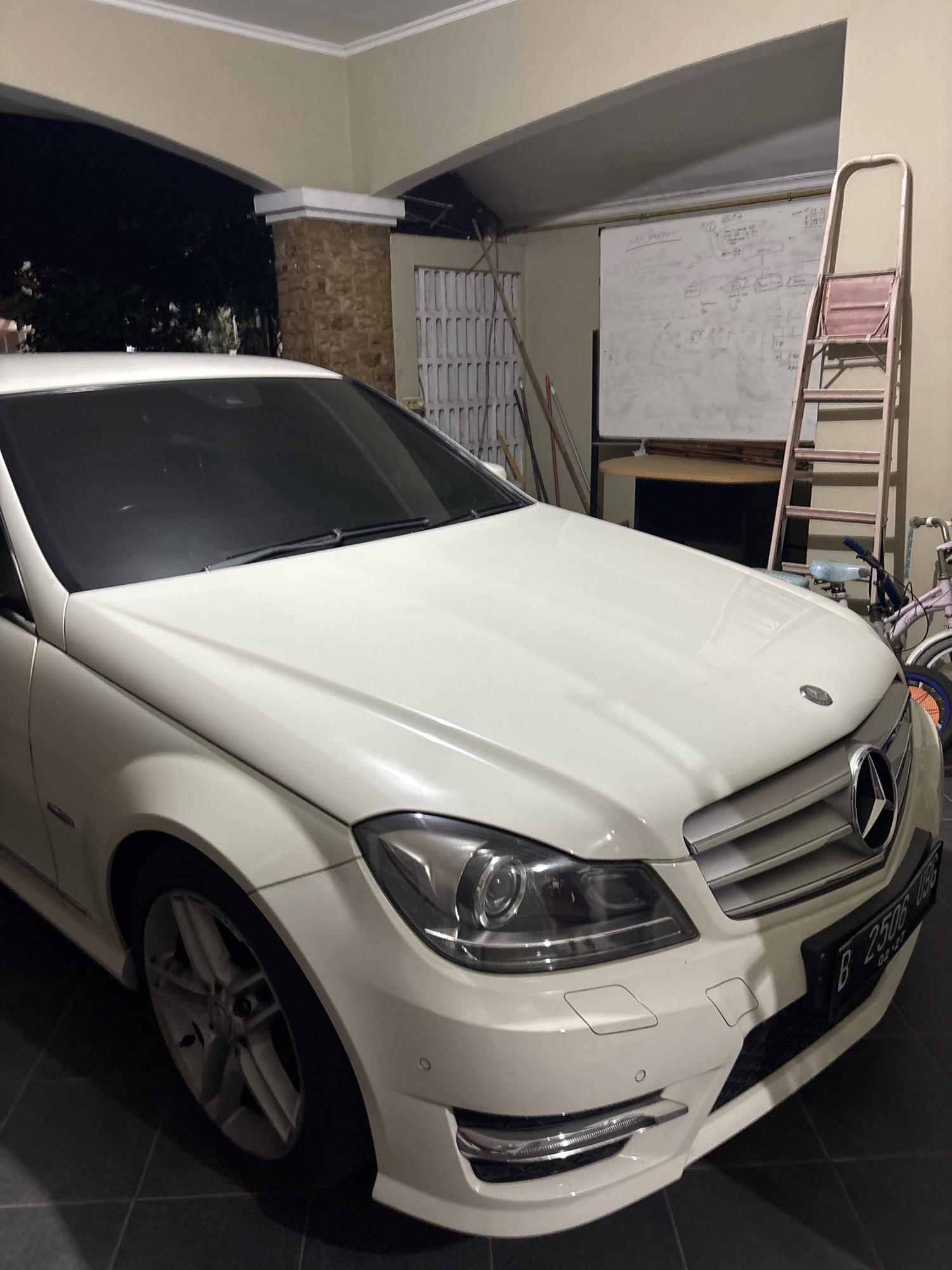 The image captures a white Mercedes-Benz C-Class sedan parked in an indoor garage or carport. The car is angled, showing its front left quarter. The license plate, B 2506 USG, is visible on the front bumper. To the right of the car, a pink step ladder leans against a whiteboard that is covered in writing. Behind the ladder, a bicycle with a pink frame and blue handlebars is parked. The garage floor is tiled in a dark color. The environment suggests a residential setting in Bekasi, Indonesia, possibly a home garage. The lighting is dim, indicating it is likely nighttime or the area is not well-lit.