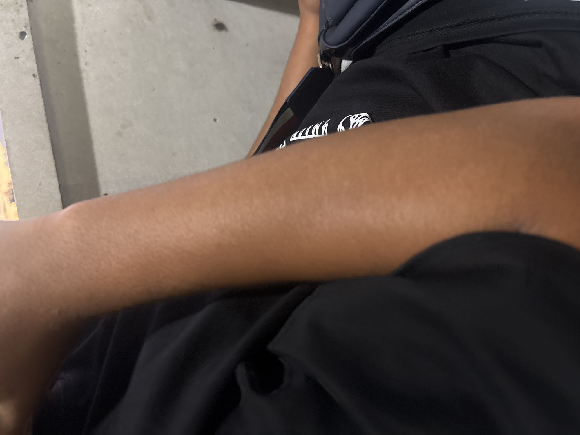 The media file captures an overhead view of a person seated in what appears to be a public space in Yerevan, Armenia. The primary subject is a person, whose right arm and upper torso are visible. The person has a medium to dark skin tone and is wearing a black short-sleeved t-shirt. On the chest of the t-shirt, there is a prominent white graphic design. This design appears stylized and abstract, possibly depicting a skeletal motif or intricate lettering, with discernible jagged lines and shapes that hint at an alphanumeric character like a 'W' or 'M'.
Draped over the person's shoulder is a dark-colored (possibly navy blue or black) shoulder bag or cross-body bag with some metallic hardware. A dark-edged object, potentially a smartphone, is partially visible, tucked near the bag or clothing.
The setting consists of a light grey, somewhat textured background that resembles a concrete or paved surface, suggesting an outdoor urban environment like a street, sidewalk, or plaza. Darker specks and stains are scattered across this surface. The lighting is even, without harsh shadows, which might indicate an overcast day or an indoor area with diffused light. No explicit activity is taking place beyond the person being at rest. No specific landmarks of Yerevan are visible, but the general context points to an everyday scene within the city. No legible text is visible other than the abstract graphic on the shirt.
