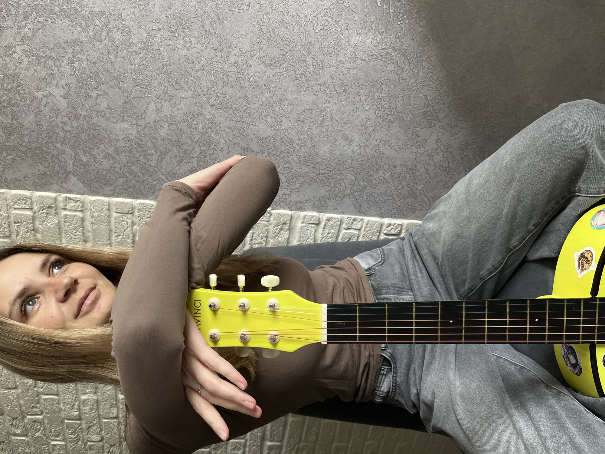A young adult female is seated indoors in the city of Chaikovsky, Russia. She has light brown hair and blue eyes, and is wearing a long-sleeved brown top and grey jeans. Her arms are crossed and resting on the headstock of a bright yellow acoustic guitar. The guitar's headstock, marked "DAVINCI," has six strings and white tuning pegs. The dark fretboard is visible, extending down toward the partially obscured, sticker-adorned yellow guitar body. The woman's left hand, resting on the guitar headstock, displays a ring on her fourth finger. She is looking upwards and to her right, away from the camera. The background consists of a white faux brick wall on the left and a textured grey wall on the right. A dark grey chair back is partially visible behind her.