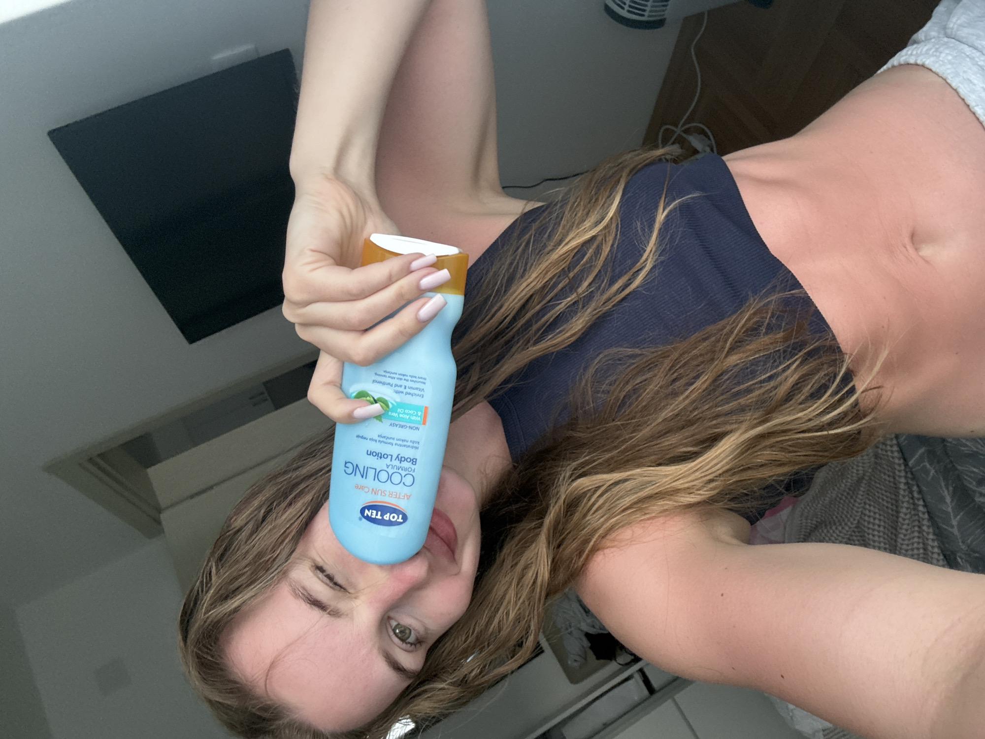 The image, rotated approximately 90 degrees clockwise, captures a young woman in an indoor setting, likely in Budva, Montenegro. She is playfully posing with a light blue bottle of "TOP TEN AFTER SUN Care COOLING FORMULA Body Lotion," holding it close to her face, touching her nose and mouth. She winks with her left eye and has a slight smile, conveying a relaxed and jovial mood.
The woman has long, wavy brown hair. She is wearing a dark blue sleeveless top and light grey shorts, with her tanned midriff visible. Her fingernails are painted with light pink polish. In the background, elements of a room are visible, including a dark rectangular object (possibly a screen or shelf) and a white fan.
The label on the lotion bottle is prominent and readable. It states "TOP TEN AFTER SUN Care COOLING FORMULA Body Lotion," emphasizing its purpose. Further details visible on the label include "Hidratantna formula koja neguje kožu nakon sunčanja" (Hydrating formula that nourishes the skin after tanning), "NON-GREASY," and "With Aloe Vera & Coco Oil." It also mentions being "Enriched with: Vitamin E and Panthenol," and that it "Nourishes the skin After tanning" and "Hrani kožu nakon sunčanje." The presence of after-sun care lotion suggests a warm climate, consistent with the specified location of Budva, Montenegro.