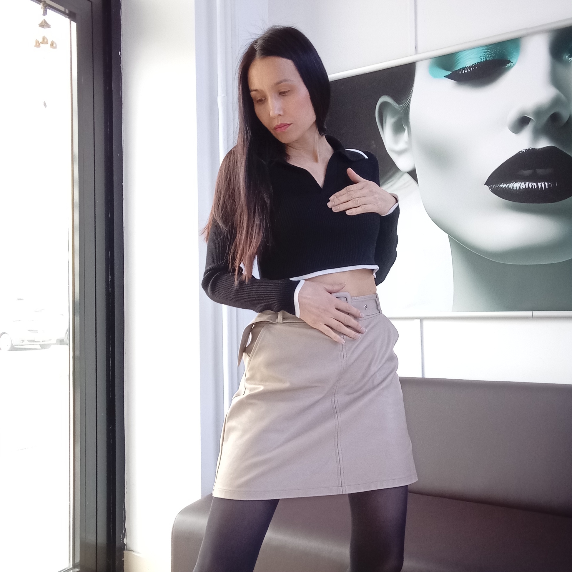 The image depicts a woman in a black, long-sleeved crop top with white trim and a beige skirt over dark tights. She is standing indoors, posing with one hand on her chest and the other on her stomach. The background includes a window with a view of a city street and a large, stylized portrait of a woman's face with dark lipstick and blue eyeshadow. The location appears to be Izhevsk, Russia.