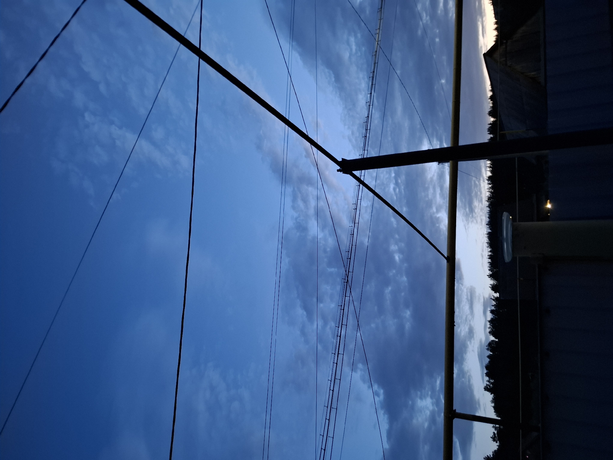 The image depicts a view of the sky with power lines and a utility pole in the foreground, possibly located in the city of Амзя, Russia. The sky is a gradient of blue hues, with some clouds visible. The power lines are dark and extend across the frame. A utility pole is present on the right side of the image, appearing metallic and somewhat obscured by shadows. In the background, there's a suggestion of trees and a building or structure, all under dark conditions that suggest evening or twilight.