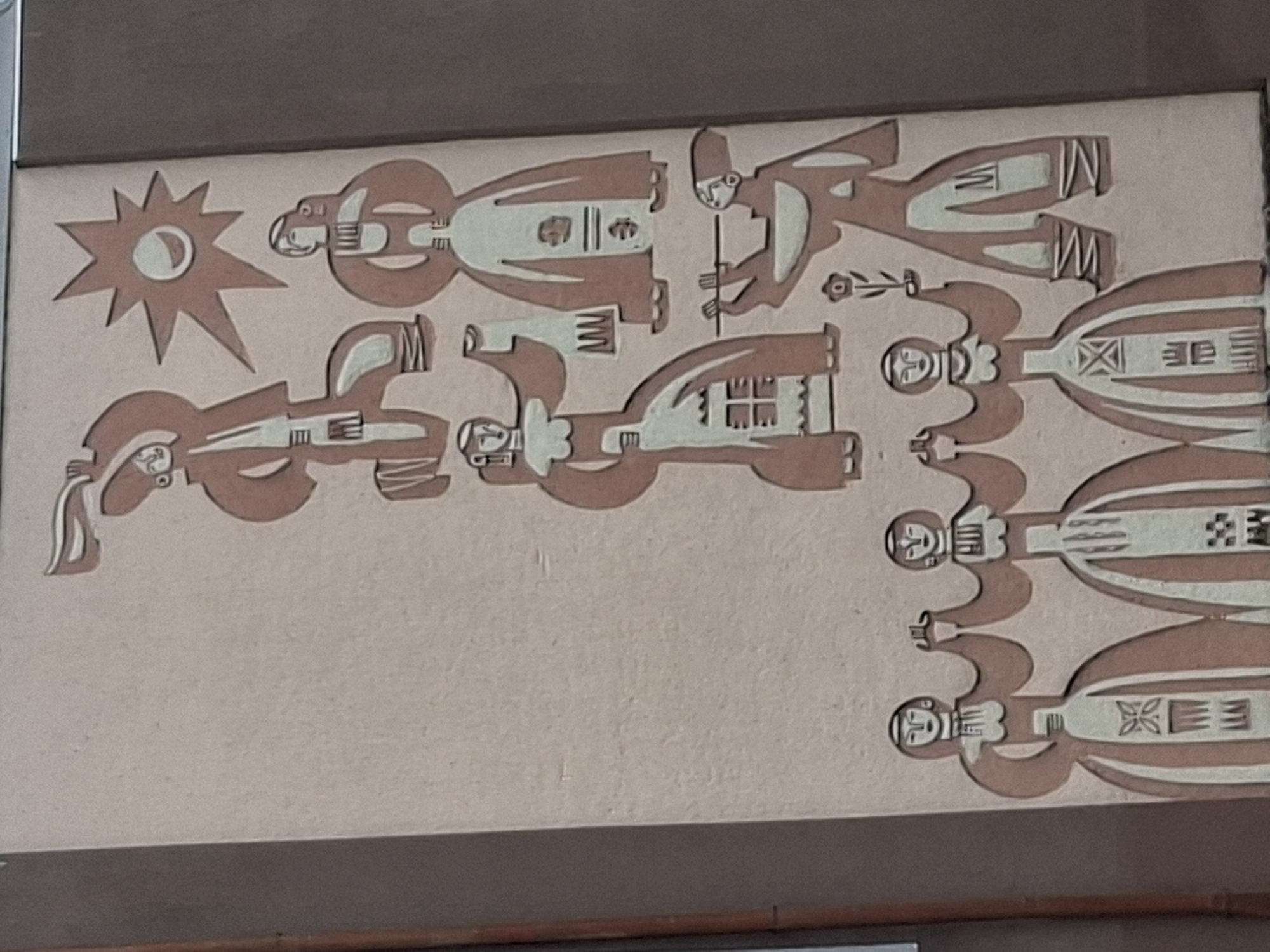 A rectangular mural depicts stylized figures in a horizontal arrangement. The mural is beige with light brown figures. On the left, a sun with rays emanates from the corner. The figures appear to be people dressed in traditional clothing, with some holding items like a flower and a staff. The figures are arranged in two rows, one above the other. The details on the clothing and the figures themselves are abstract and geometric. The location is Galabovo, Bulgaria.