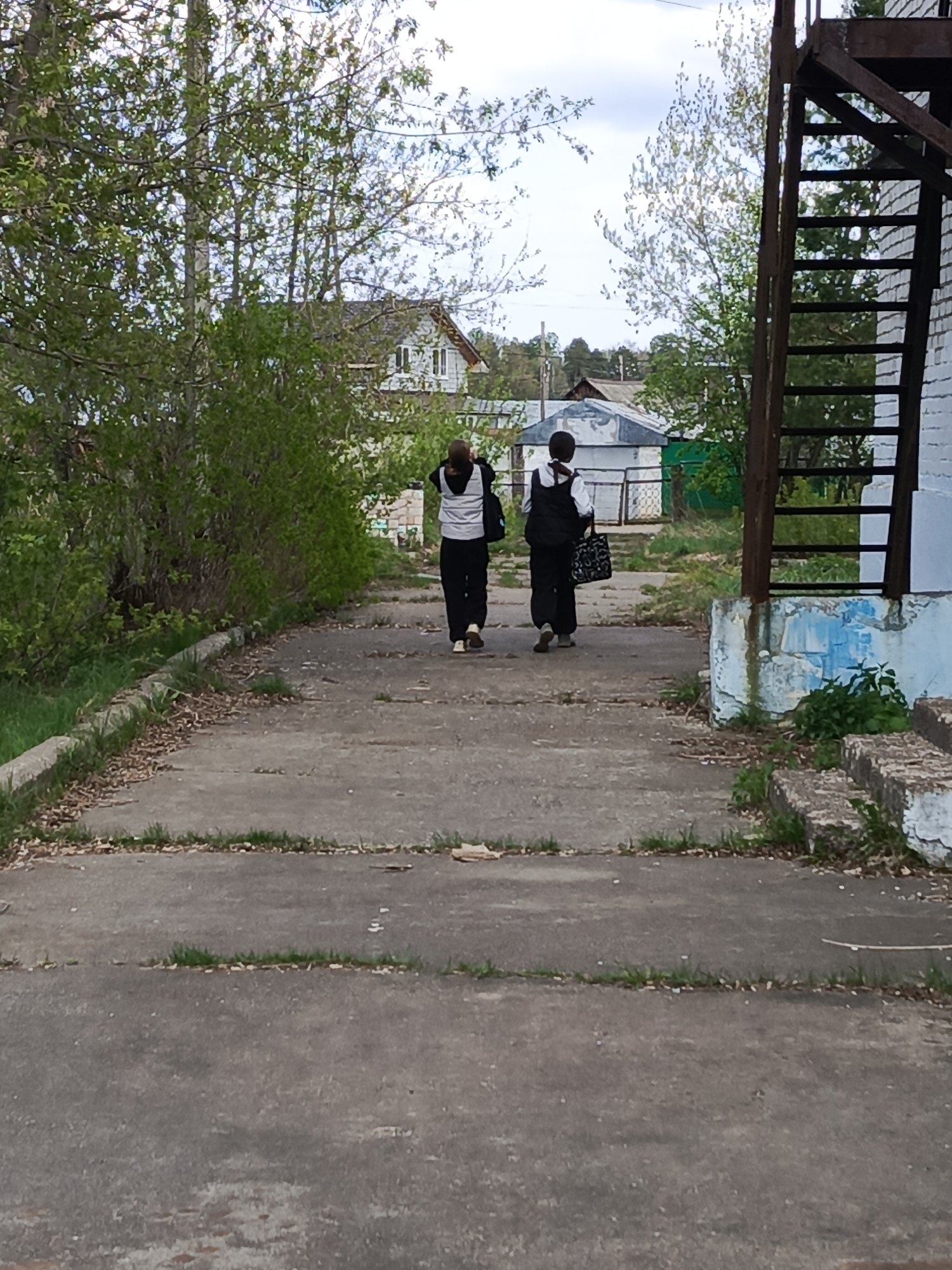 Two individuals walk away from the viewer on a cracked concrete path in a townscape in Russia. To the left is green foliage, and to the right is a weathered building with a rusty fire escape ladder attached. In the background, various houses and structures are visible. The city is Chaikovsky, Russia.