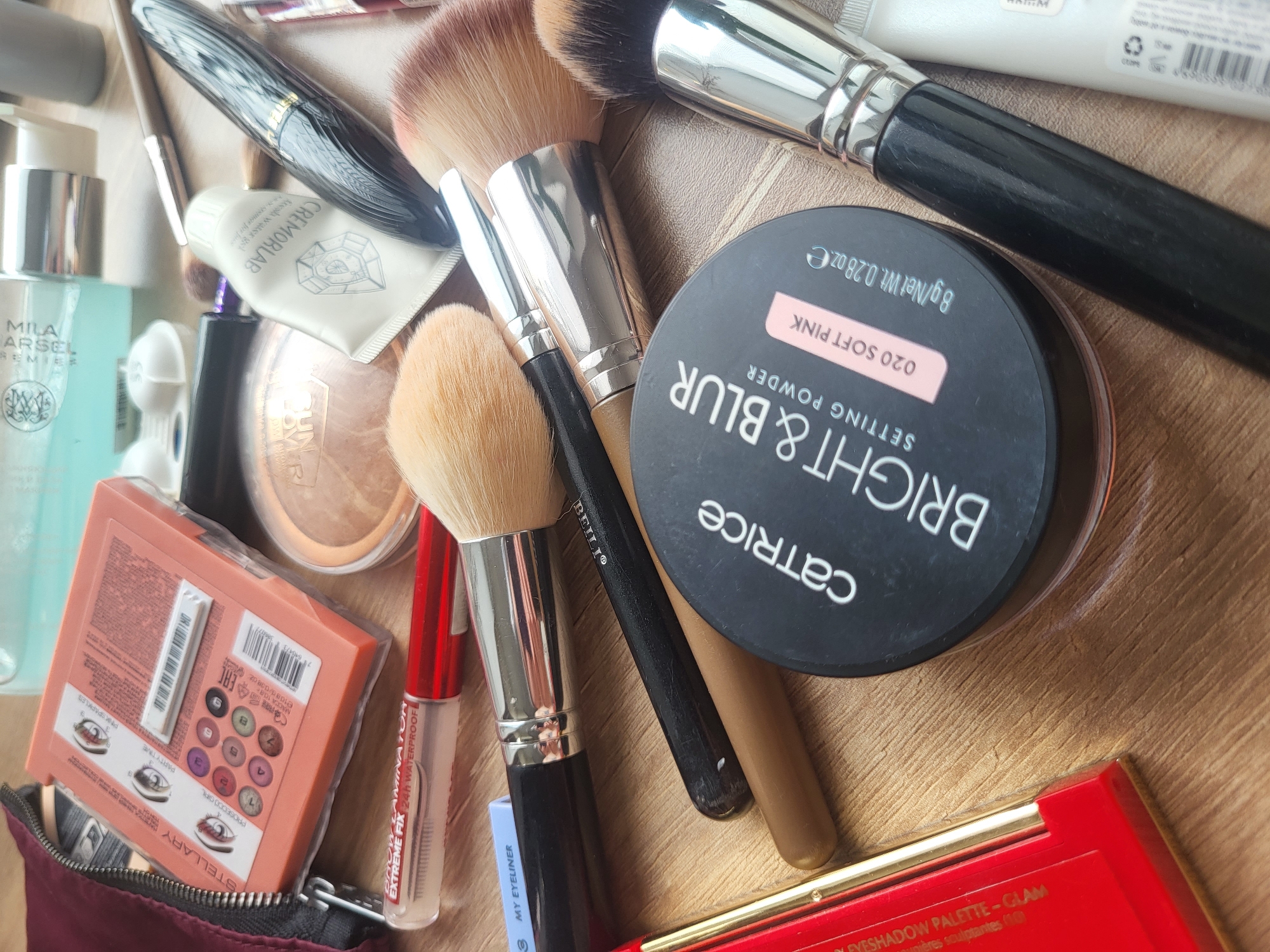 The image shows a collection of makeup products and tools arranged on a light brown surface. Prominent items include Catrice Bright & Blur Setting Powder (020 Soft Pink), a Stellary eyeshadow palette, makeup brushes of varying sizes, Mila Arsel Premier skincare, Cremorlab skincare, and other makeup items. The labels indicate brands such as "BEILI" on a brush, "MY EYELINER", and "HOLYNIN EXTREME FIX." The Stellary palette packaging displays the names "PROSECCO GIRL," "PARTY TIME," and "PINK SPARKLES." Given the presence of the Stellary and other brands with labels in Cyrillic script, the image might originate from Russia. However, due to the generic nature of the makeup items and the lack of distinct landmarks or identifiable elements in the background, it is impossible to conclusively determine if the location is specifically Chaikovsky, Russia.