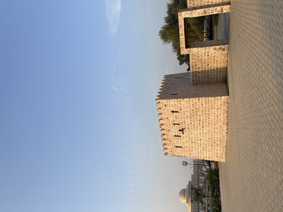The image depicts a historical stone fort structure in Sharjah, United Arab Emirates, specifically located within the Sharjah Heritage Area, near Bank Street. The central feature is a light-colored, square-plan, stone-built fort with a crenellated parapet roofline and several small, rectangular openings (embrasures) on its visible faces. Attached to the fort's right side is a lower stone wall segment, which leads to a freestanding stone archway providing access to an area beyond. The foreground consists of a large, light-grey paved plaza, constructed with interlocking pavers. In the distant left background, a modern building with a prominent domed roof and multiple stories is visible, along with green landscaping and palm trees. To the right of the fort and through the archway, dense green foliage from trees is present. The sky above is clear and light blue with minimal, wispy clouds. No individuals or vehicles are present within the frame. The scene is illuminated by natural light, casting soft shadows.