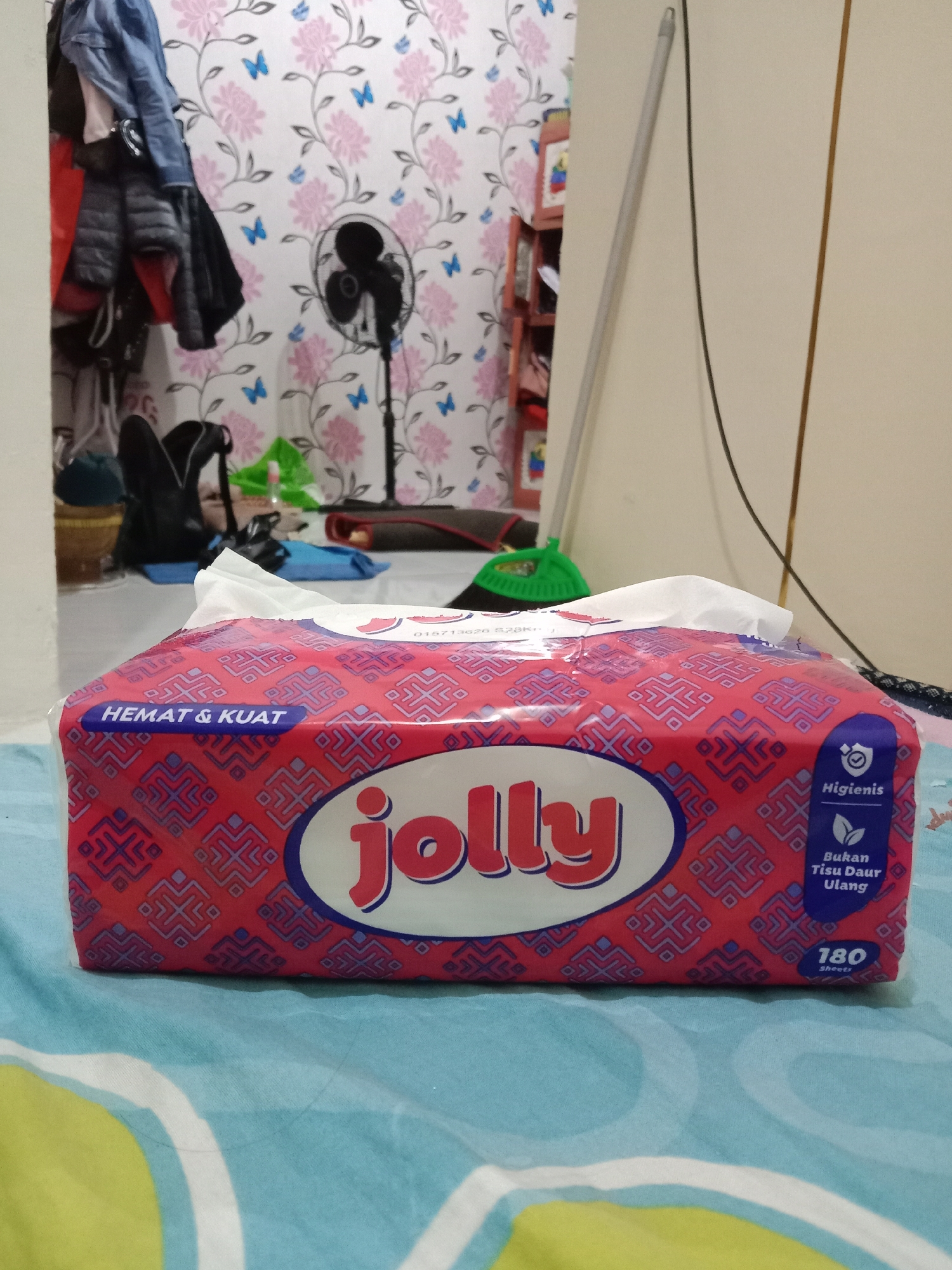 This is a medium-shot, eye-level, horizontal image of a red and blue patterned tissue box labeled "jolly" placed on a blue and green patterned fabric surface. The tissue box is centered in the frame and positioned towards the bottom. The packaging features Indonesian text: "HEMAT & KUAT" (economical & strong) on the left and "Hijienis" (hygienic) and "Bukan Tisu Daur Ulang" (not recycled tissue) on the right. A small numeral "180 Sheets" is visible at the bottom right corner of the box.

The background shows a wall with floral wallpaper in pink and white with blue butterfly accents. To the left, a dark object, possibly a shelf or wardrobe, is partially visible with clothes hanging on it. A black oscillating fan is positioned against the wallpaper. To the right of the fan, a light gray pole leans against the wall, and a black cable hangs down. Further to the right, a cream-colored surface, likely a wall or furniture, is visible.

The location is identified as Brangsong, Indonesia. The scene appears to be an indoor setting, possibly a bedroom or living area, characterized by domestic objects and decorative wallpaper.