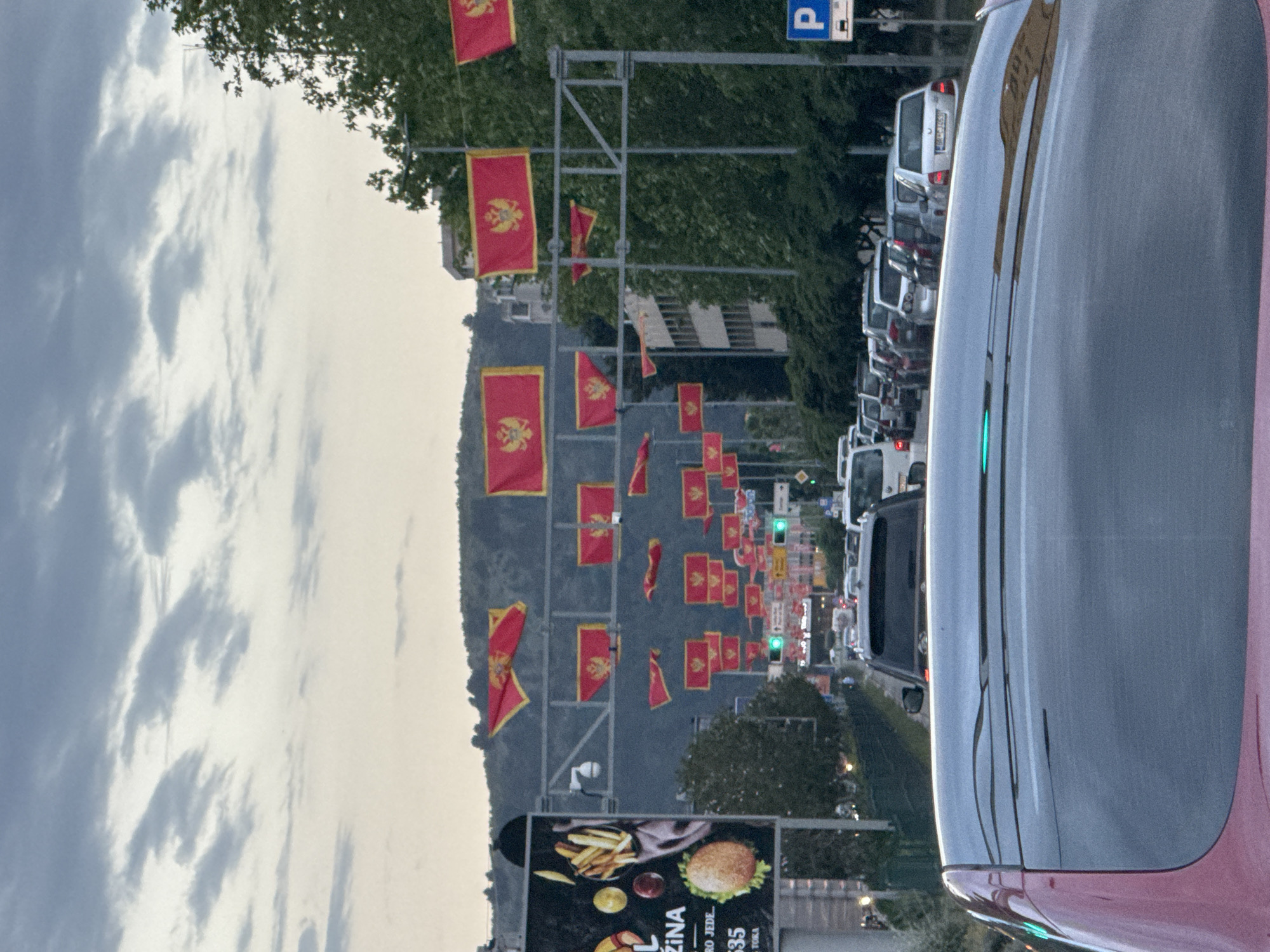 A street in Bar, Montenegro, lined with Montenegrin flags hanging from overhead supports. A row of parked cars lines the right side of the street. A bus or large vehicle is visible in the foreground on the right. An advertisement for food is on the left side of the street. The sky is overcast. Traffic lights are visible in the distance.