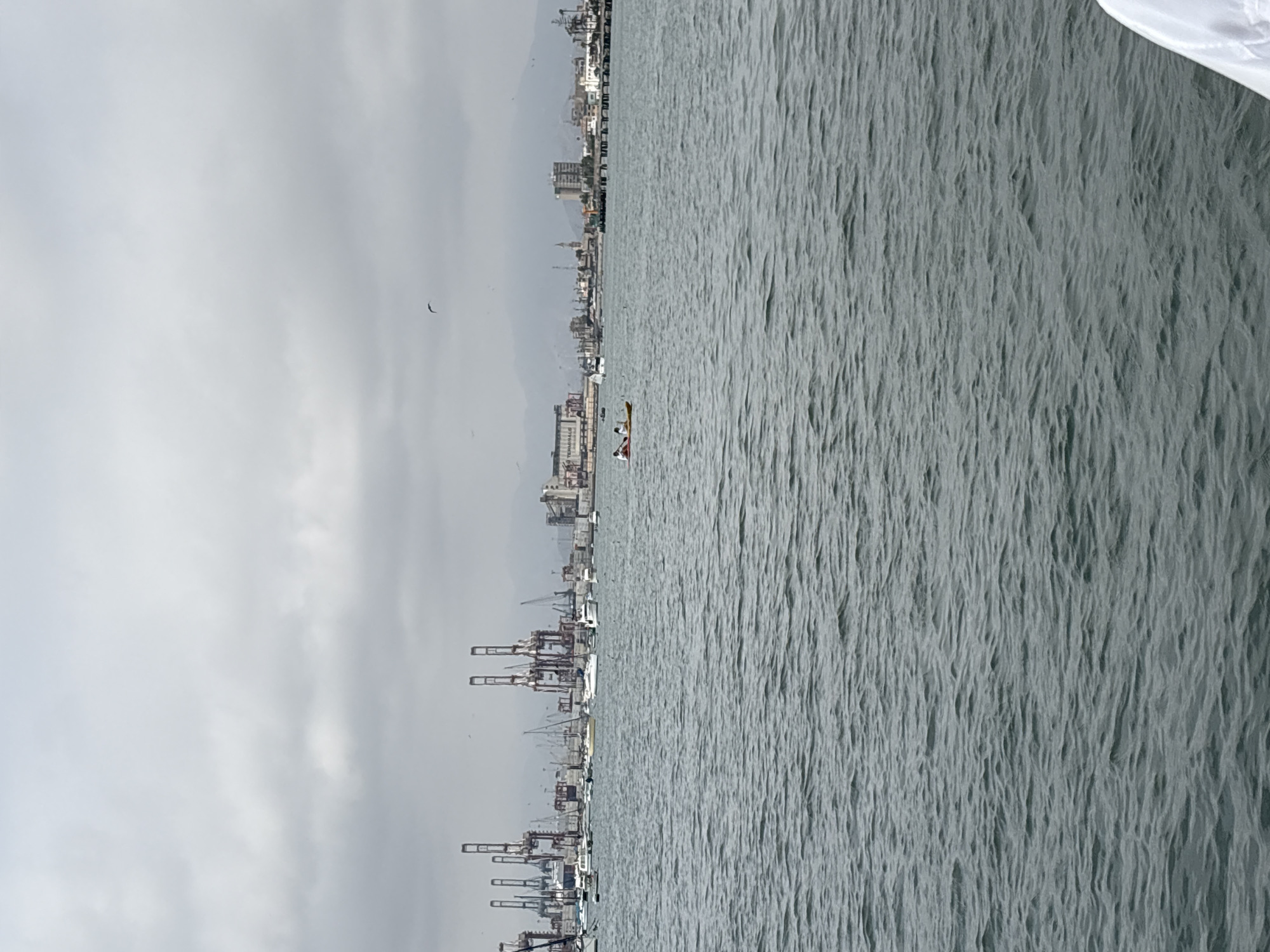 The image captures a waterfront scene in La Punta, Peru, with a body of water in the foreground and a city skyline in the background. The water is choppy, with small waves indicating a moderate breeze. Two individuals are visible in a yellow kayak, paddling on the water. In the middle ground, a variety of boats are moored, including sailboats and smaller vessels. The background is dominated by the industrial port of Callao, characterized by numerous cranes, large buildings, and shipping containers. Mountains form a distant backdrop to the urban and industrial landscape. The sky is overcast with gray clouds, suggesting a cloudy or hazy day.