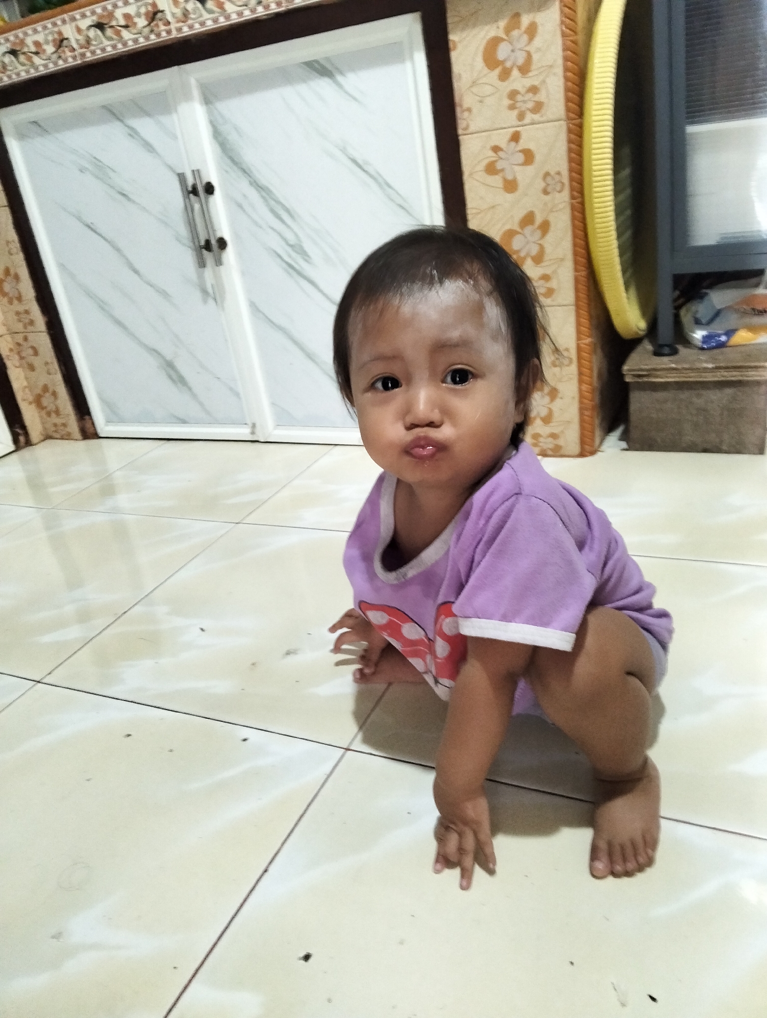A child is on a tiled floor in front of a white wardrobe with a decorative border. The child is wearing a purple t-shirt and is in a squatting position, looking at the camera with a pout. The background includes a yellow object and a brown storage box. The image appears to be taken indoors, likely in a residential setting in Muncar, Indonesia.