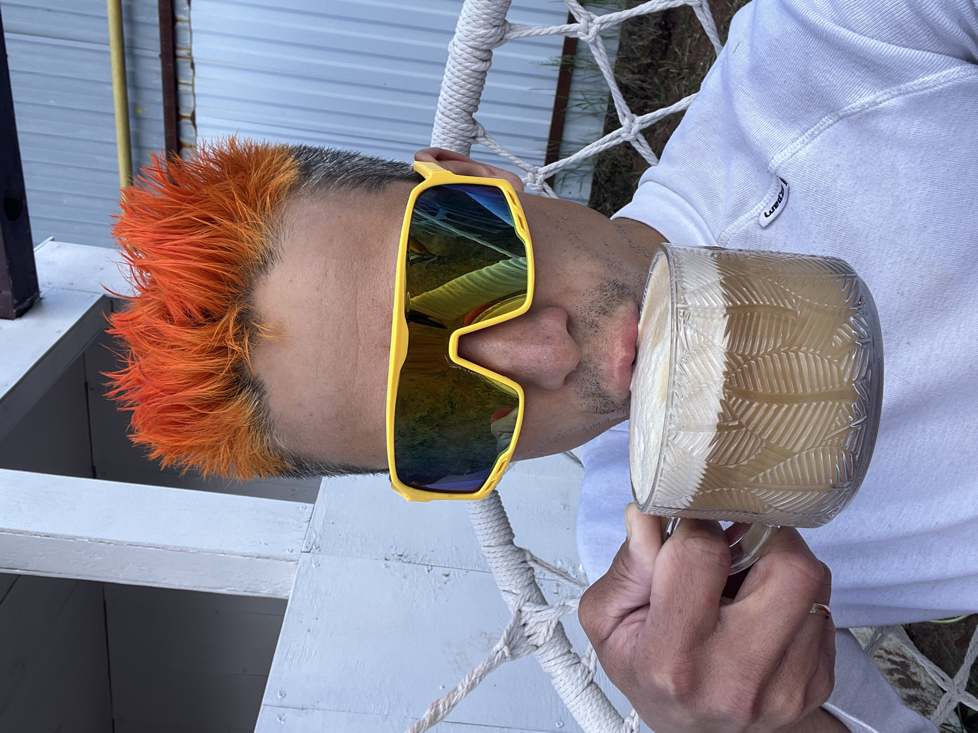 A person with orange spiky hair and yellow sunglasses is drinking from a glass mug. The person is wearing a white sweatshirt. The mug is decorated with a leaf pattern and appears to contain a milky brown liquid. The background includes a white rope hammock or swing, with white painted wood structures visible. The precise location cannot be confirmed as Chaikovsky, Russia.