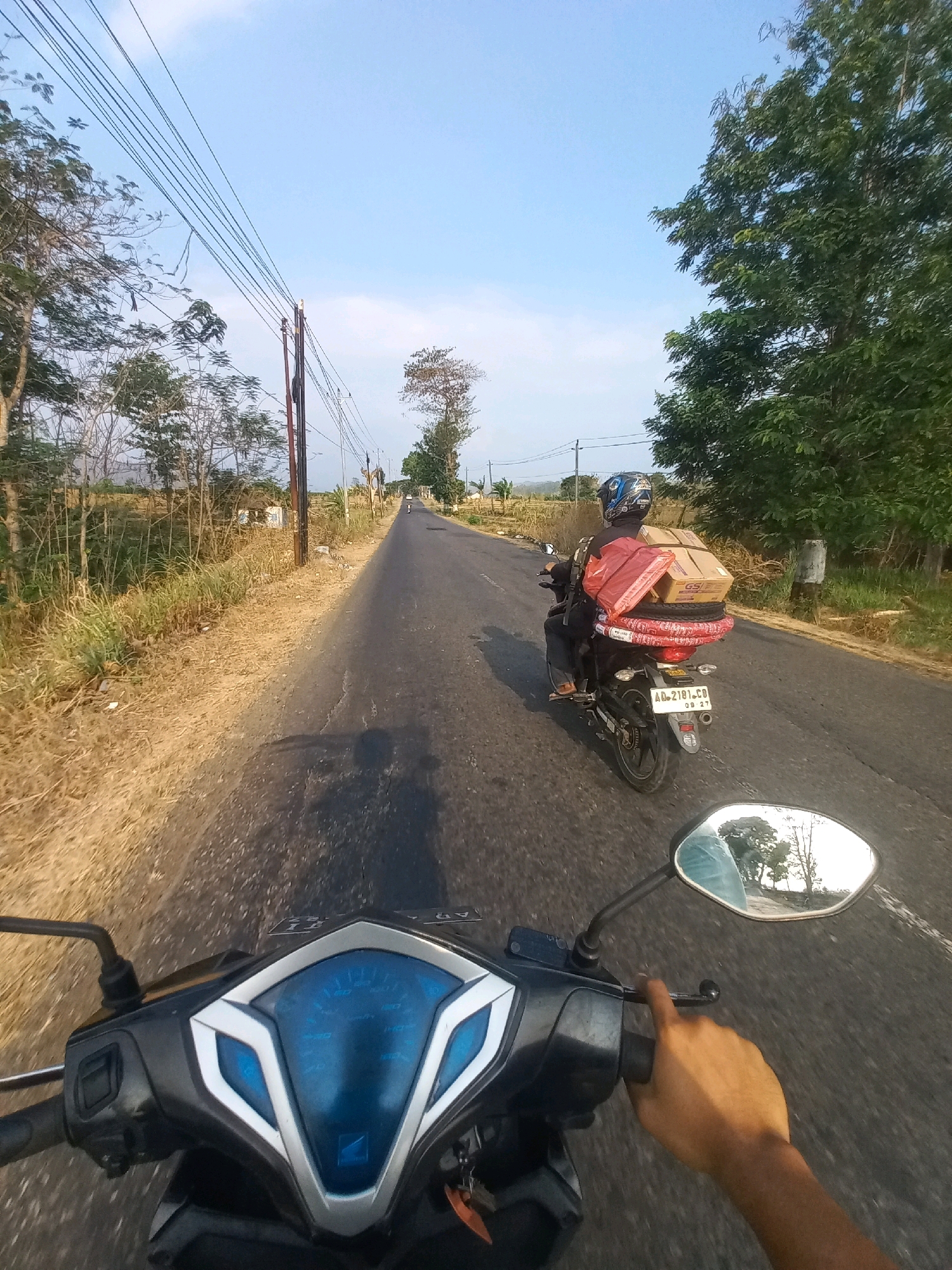 A first-person perspective shot of a person riding a motorcycle on a paved road under a clear sky. The rider's hand is visible on the handlebar, and the motorcycle's dashboard is in the foreground. The road is flanked by dry grass and trees. In the distance, another motorcycle is visible, carrying several packages and what appear to be tires on its rear rack. The rider of the second motorcycle is wearing a helmet. The scene suggests a rural or semi-rural area in Baturetno, Indonesia, likely during daylight.