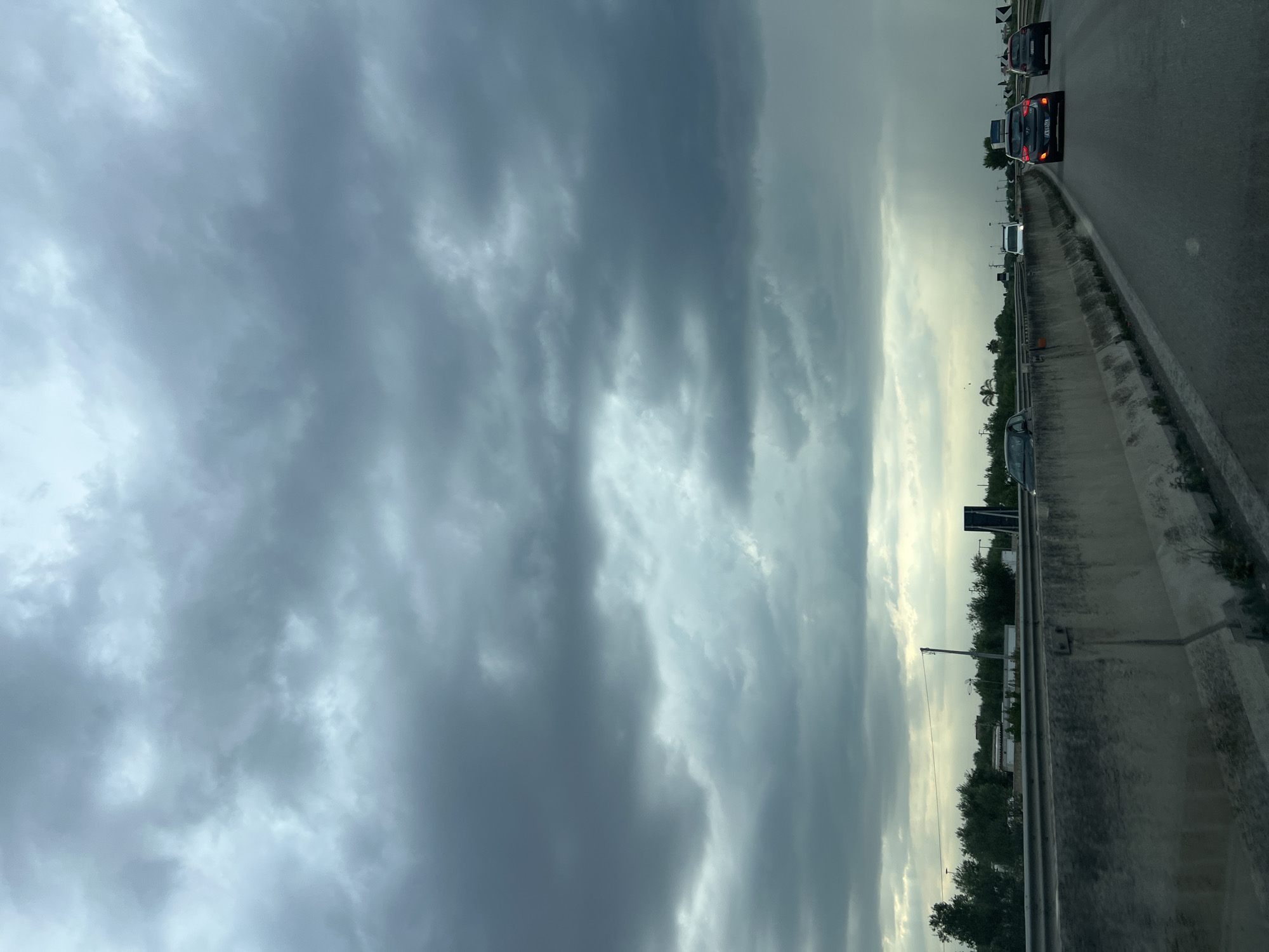 The image depicts a view from a vehicle traveling on a highway, likely the SS16bis, near Bisceglie, Italy. The sky is overcast with dark, heavy clouds, suggesting impending rain, with a visible streak of rain falling in the distance on the right side of the frame. The highway has a concrete divider separating traffic, with several cars visible on the road ahead. Trees and some buildings are present in the background beyond the divider. The road surface is asphalt.