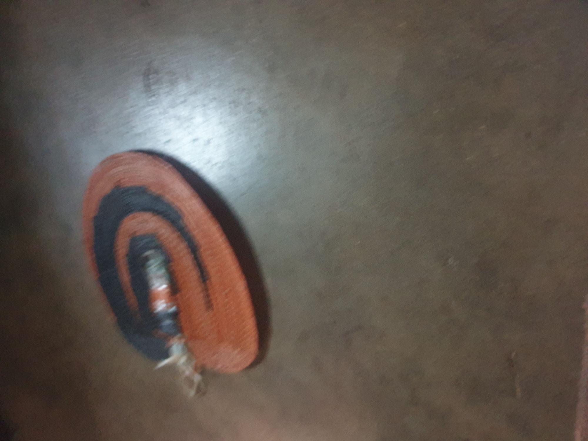 An orange and black coiled object, resembling a small rug or mat, is positioned on a textured, dark brown surface. The object has a spiral pattern, with the black material forming concentric rings within the orange base. A thin, light-colored string or cord is attached to the left side of the coiled object, appearing to be tied around it. The surface it rests on has a somewhat grainy or worn appearance, with faint lighter-colored marks visible. The lighting suggests an indoor environment, possibly dimly lit.