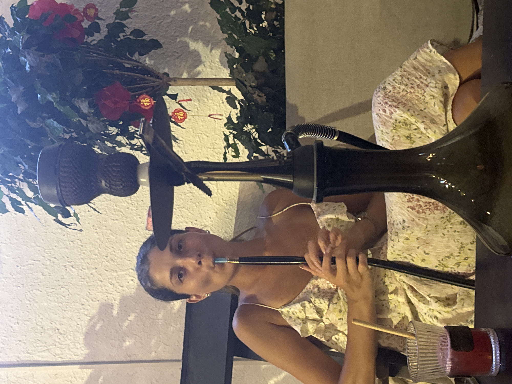 A woman is seated, smoking a hookah. She wears a floral print dress with thin straps. The hookah is black and comprises a base, a hose she holds to her mouth, and a bowl. Beside her is a glass with a straw and a dark red liquid. The location is likely outdoors given the foliage and patterned wall. The setting is likely a restaurant or lounge in an urban location (possibly Đà Nẵng, Vietnam).