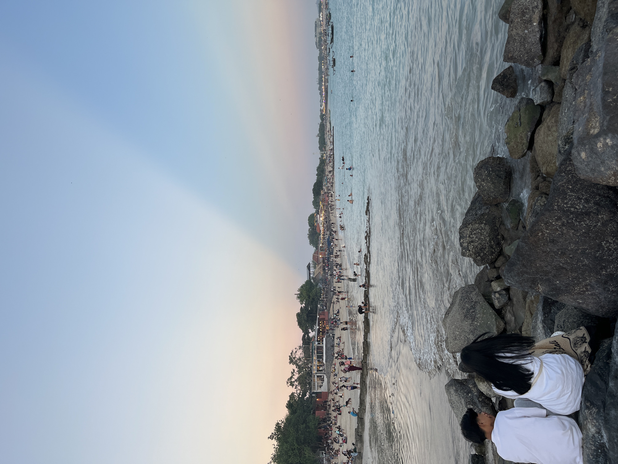 An eye-level, medium shot captures a serene beach scene in Kuta, Indonesia, during what appears to be sunset or late afternoon. The sky, a gradient of soft blue and pale pink, is dramatically bisected by a diagonal ray of sunlight, creating a striking visual effect. In the foreground, two individuals, a male and a female, are seated on dark, textured rocks, their backs to the viewer. Both are wearing white tops. The male has short dark hair, while the female has long, dark hair. They are positioned on the left side of the frame, overlooking the ocean.
The middle ground reveals a bustling beach teeming with activity. Numerous people are scattered across the sandy shore and wading in the shallow waters. Some are engaged in swimming, others are walking, and a few appear to be sunbathing. Along the coastline, buildings with illuminated windows suggest restaurants or shops, contributing to the vibrant atmosphere. Trees dot the landscape behind the beach, and several boats are visible in the distance on the water, adding to the sense of a lively coastal town.
The water itself is a calm, pale blue, with gentle waves lapping at the shore. The transition from wet sand to dry sand is clearly defined. The overall impression is one of a picturesque and lively beach setting, characteristic of popular tourist destinations like Kuta.