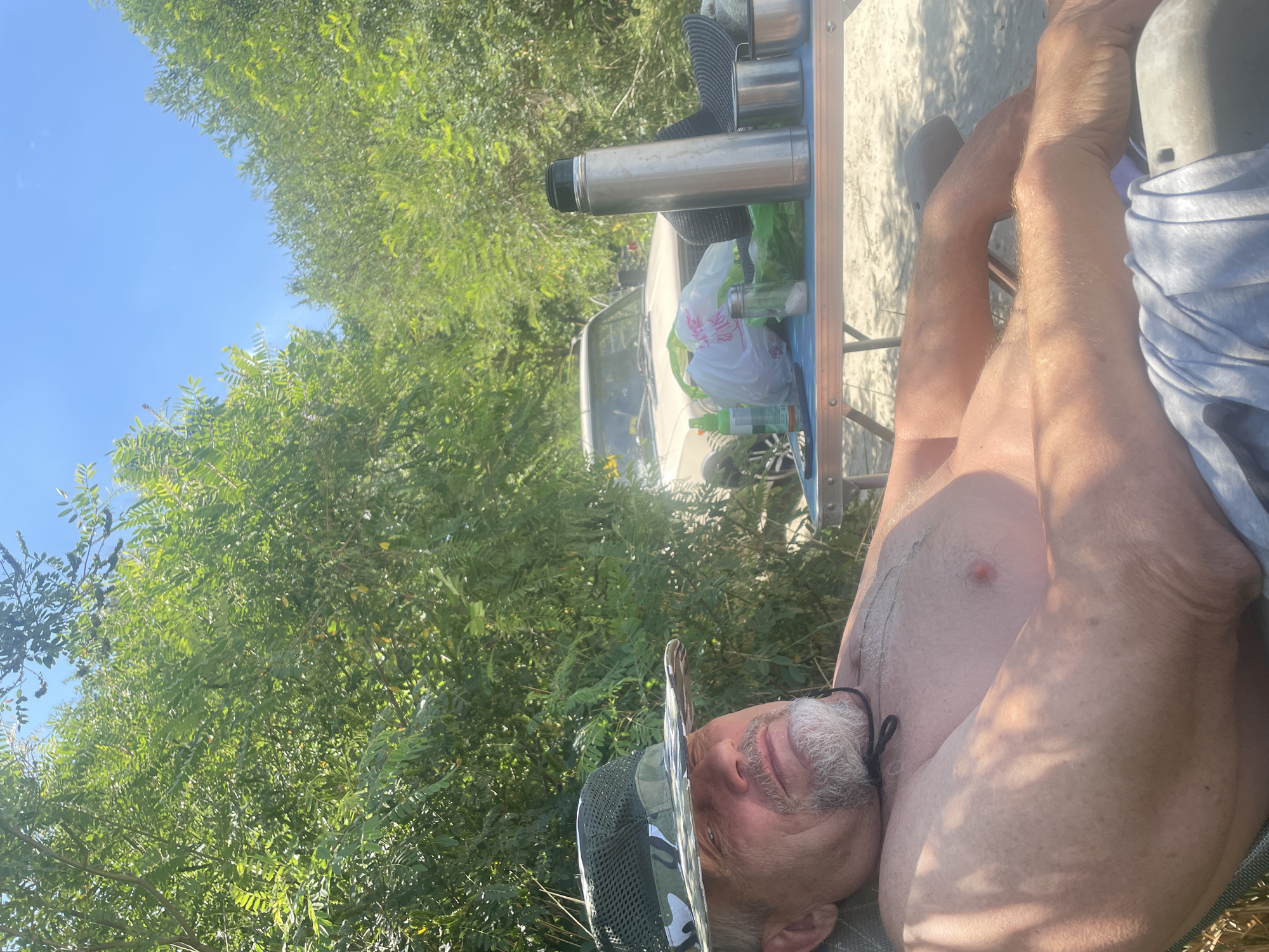 A middle-aged man wearing a camouflage hat is relaxing in a chair outdoors. He is shirtless and appears to be in a natural setting with trees and foliage surrounding him. In the background, a car is parked, and a table is set up with various items, including a thermos, cups, and bags. The weather appears sunny with a clear blue sky. The scene suggests a recreational outing, possibly a picnic or camping trip. The location is identified as Azov, Russia.