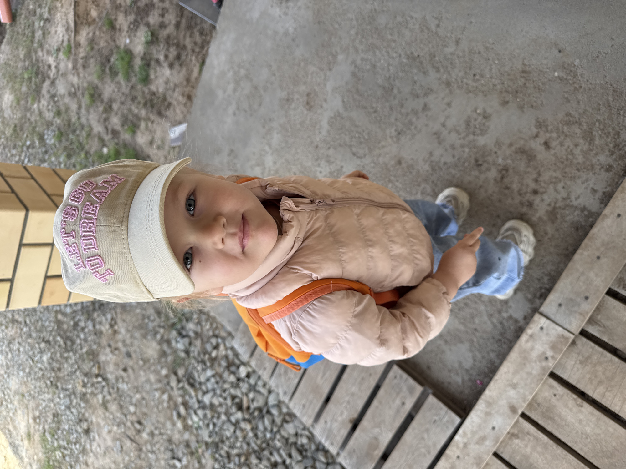 A young child with light skin, blue eyes, and light hair is standing outdoors. She is wearing a light pink quilted jacket over an orange and blue backpack, blue jeans, and light-colored sneakers. On her head is a light tan baseball cap with pink lettering that reads "LET'S GO DREAM." The child is pointing with her right index finger.
The ground surface around the child consists of gray concrete, gravel, and wooden planks. A brick structure is visible at the edge of the frame.
The location is Ферма, Russia.
