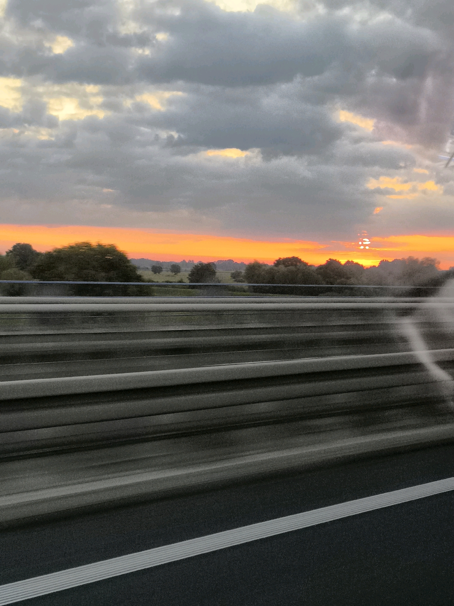 The image depicts a sunset over a rural landscape, viewed from a moving vehicle. The sky is filled with dramatic clouds, with a vibrant orange and yellow glow emanating from the setting sun on the horizon. Silhouetted trees and fields are visible in the distance. In the foreground, a metal guardrail is blurred due to motion, suggesting the perspective of a passenger. The road surface is dark asphalt with white markings. The image appears to have been taken in or near Dąbrowica, Poland, as indicated by the metadata.